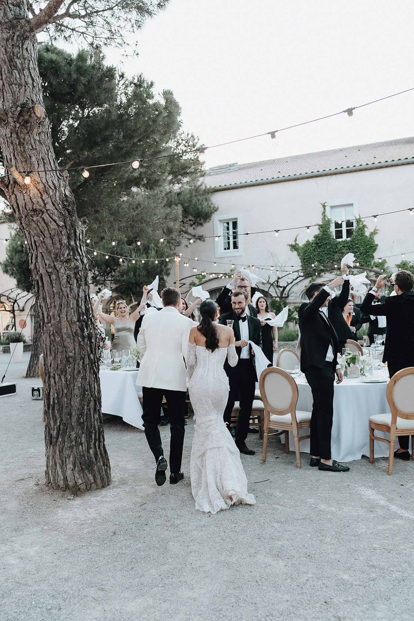 Couple entering outdoor reception as guests wave white napkins under string lights at Provencal mas