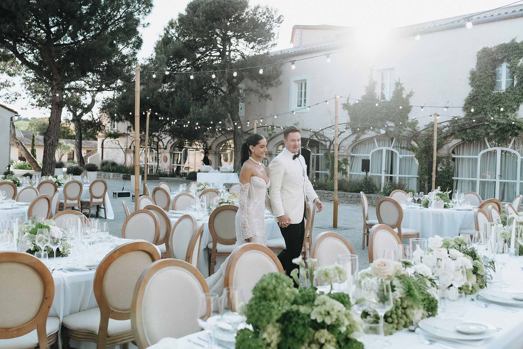 Bride in lace gown and groom in white dinner jacket walking through courtyard reception with round tables