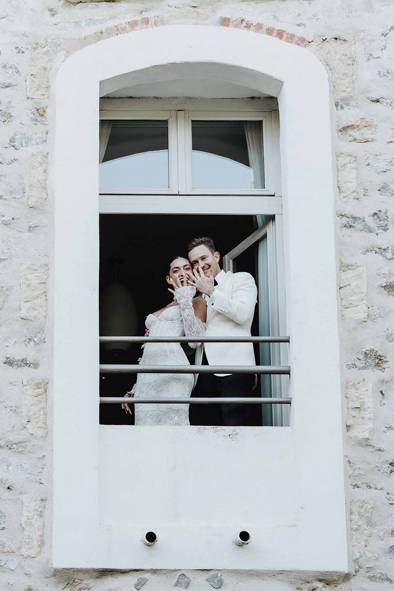 Couple laughing in arched window of limestone chateau bride in lace gown groom in white dinner jacket