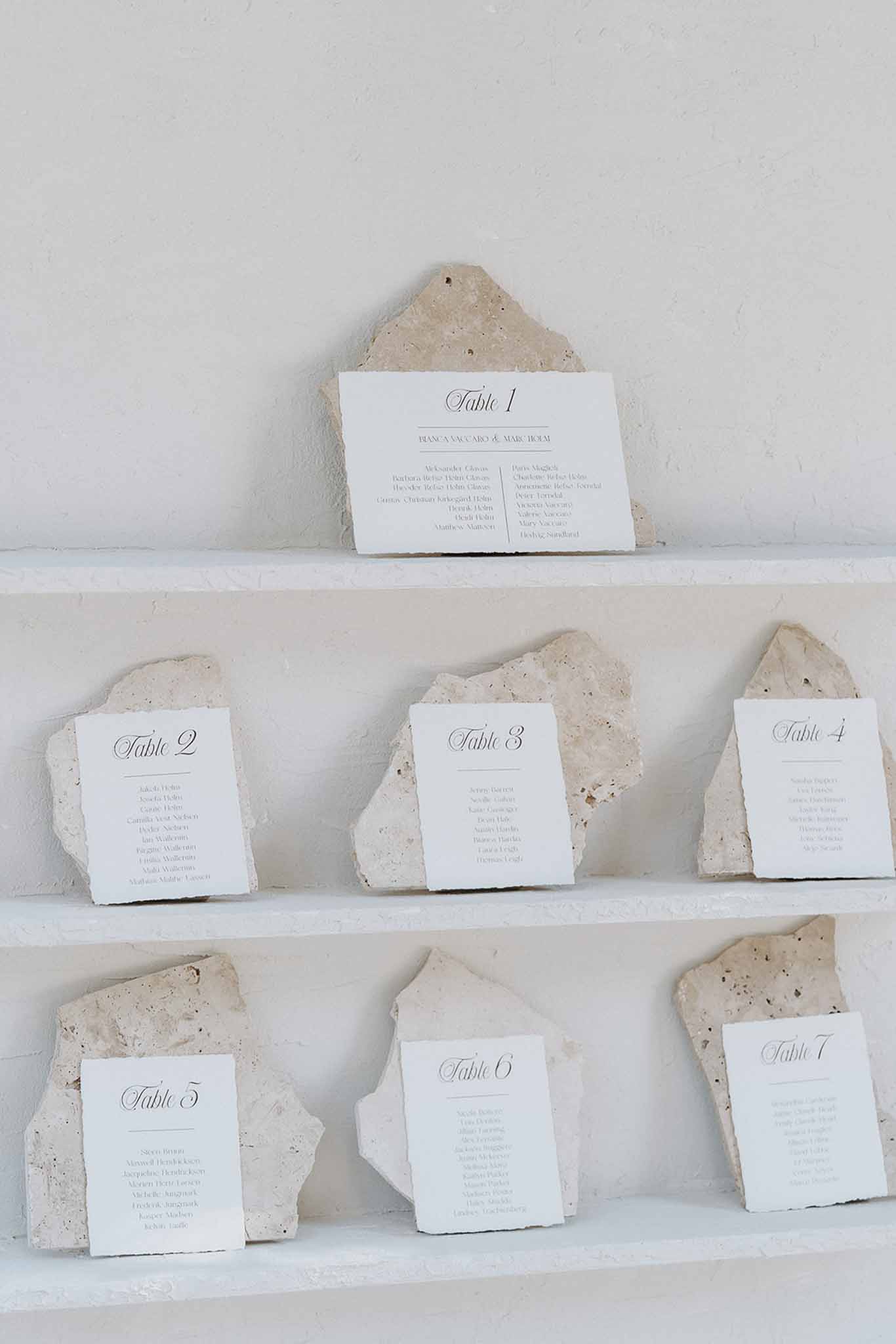 Seven table seating cards in travertine stone holders on white shelves in minimal modern display