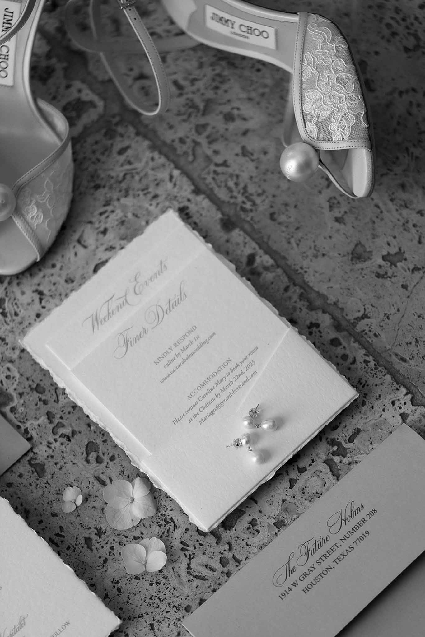 Black-and-white flat lay of wedding stationery, pearl earrings, and Jimmy Choo lace heels on stone surface