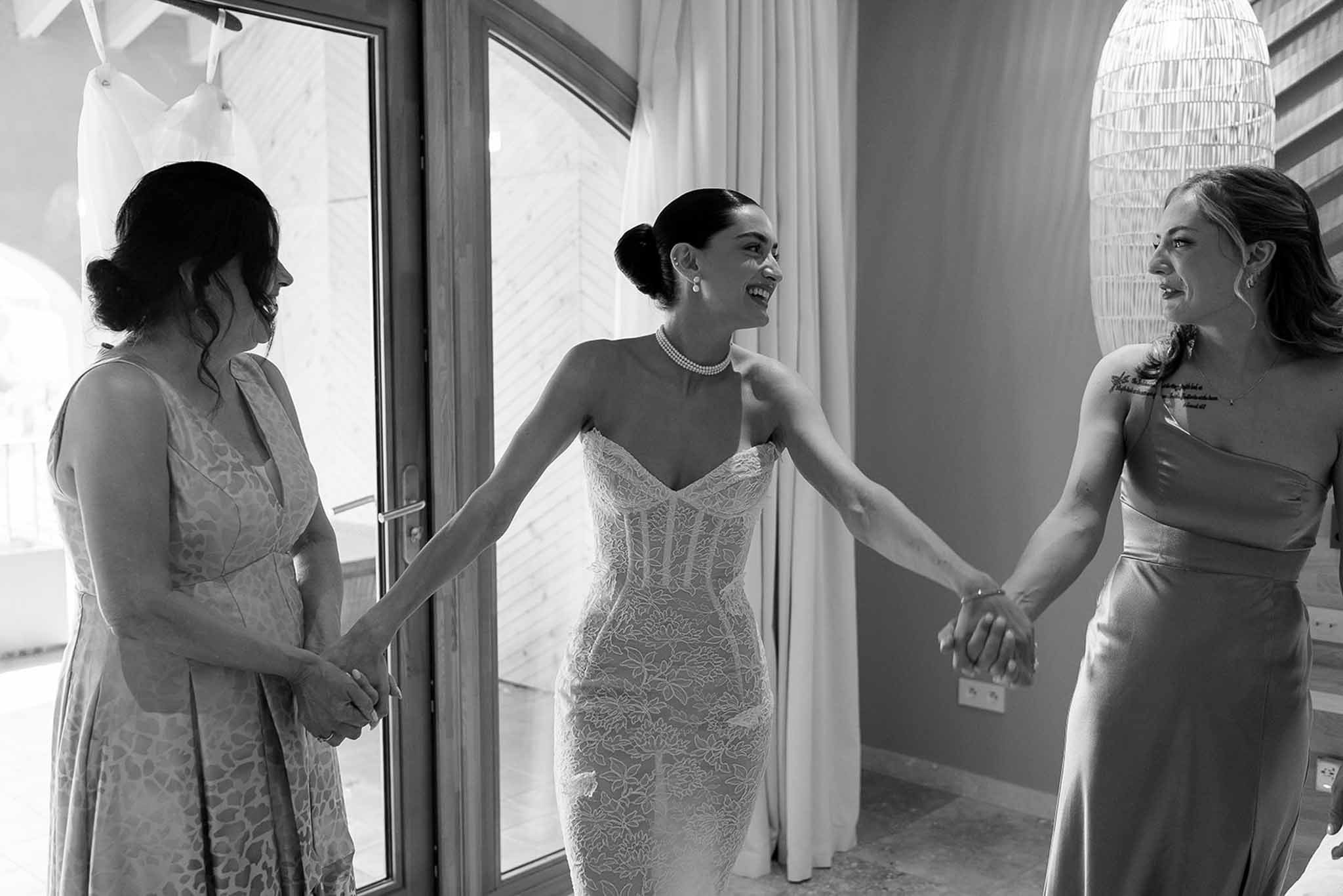 Black and white bride in strapless lace gown with pearl choker holding hands with two laughing women at glass doors