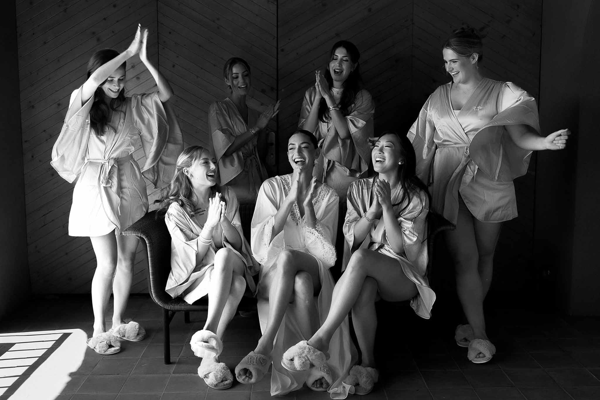 Black and white photo of bride and six bridesmaids laughing together in matching satin robes