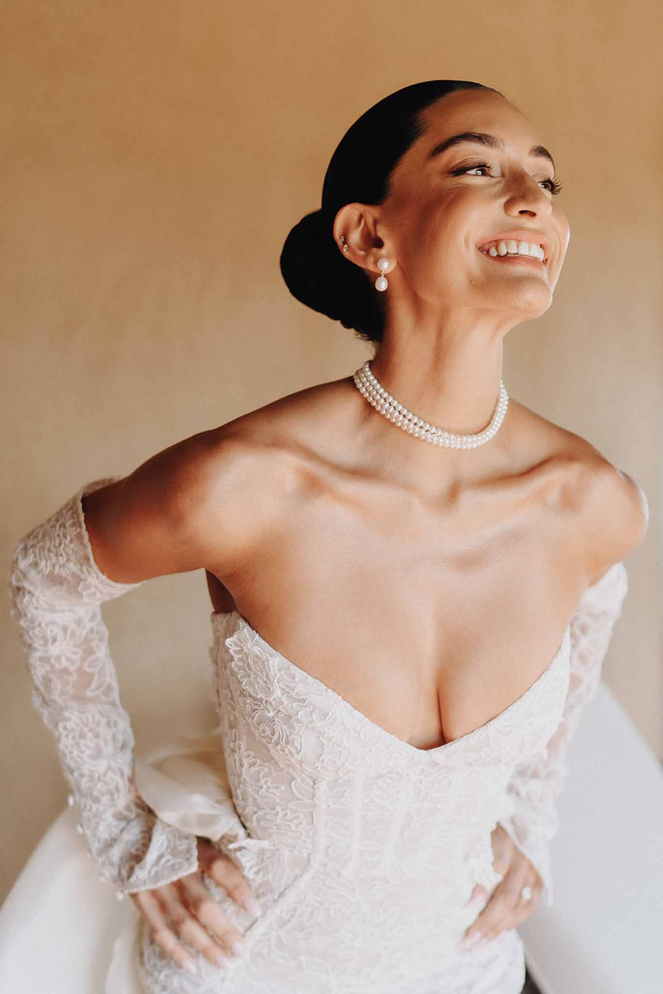 Bride smiling in ivory lace V-neck dress with off-the-shoulder lace gloves and pearl choker against ochre wall