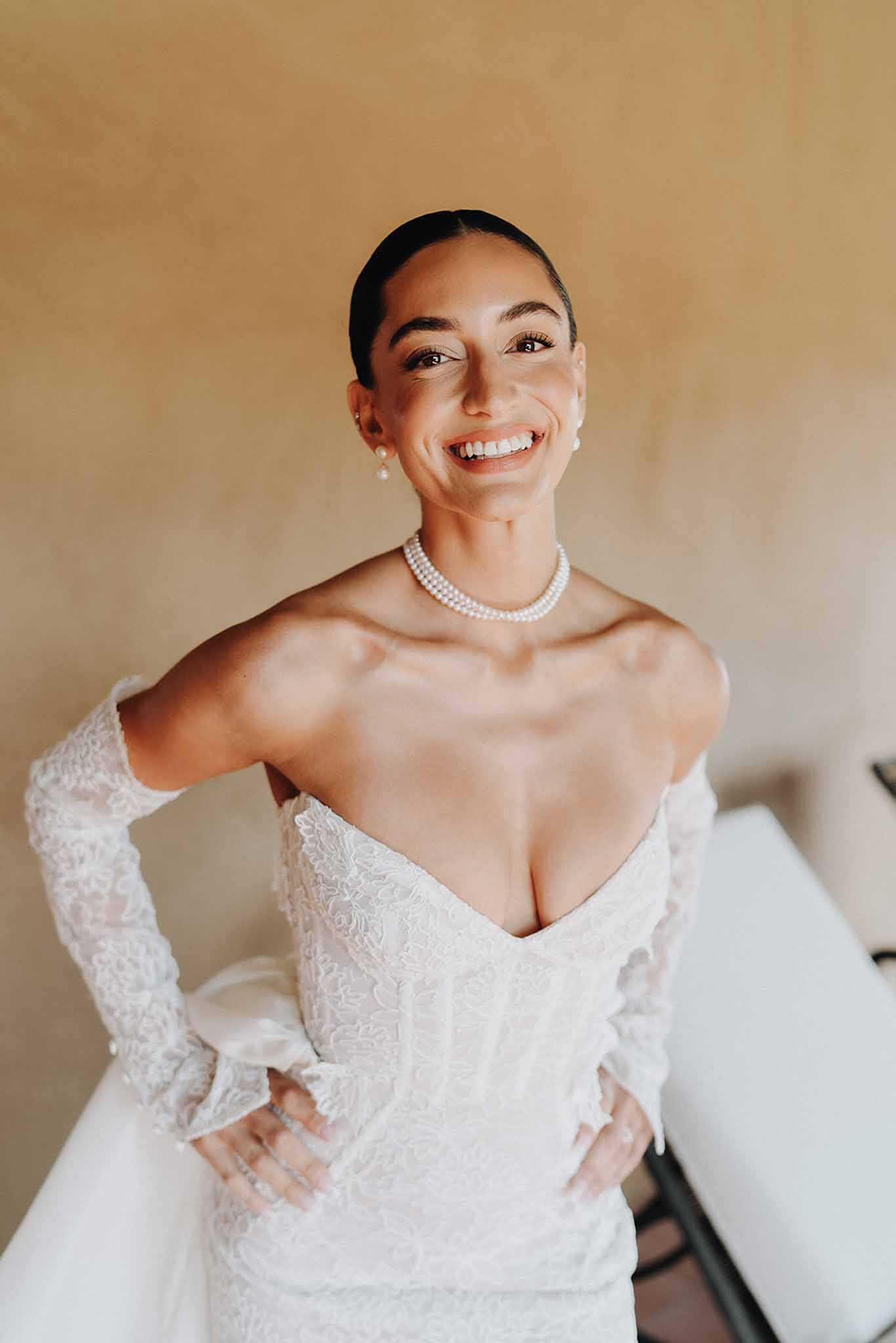 Smiling bride in off-shoulder lace gown with pearl choker and lace gloves against ochre wall