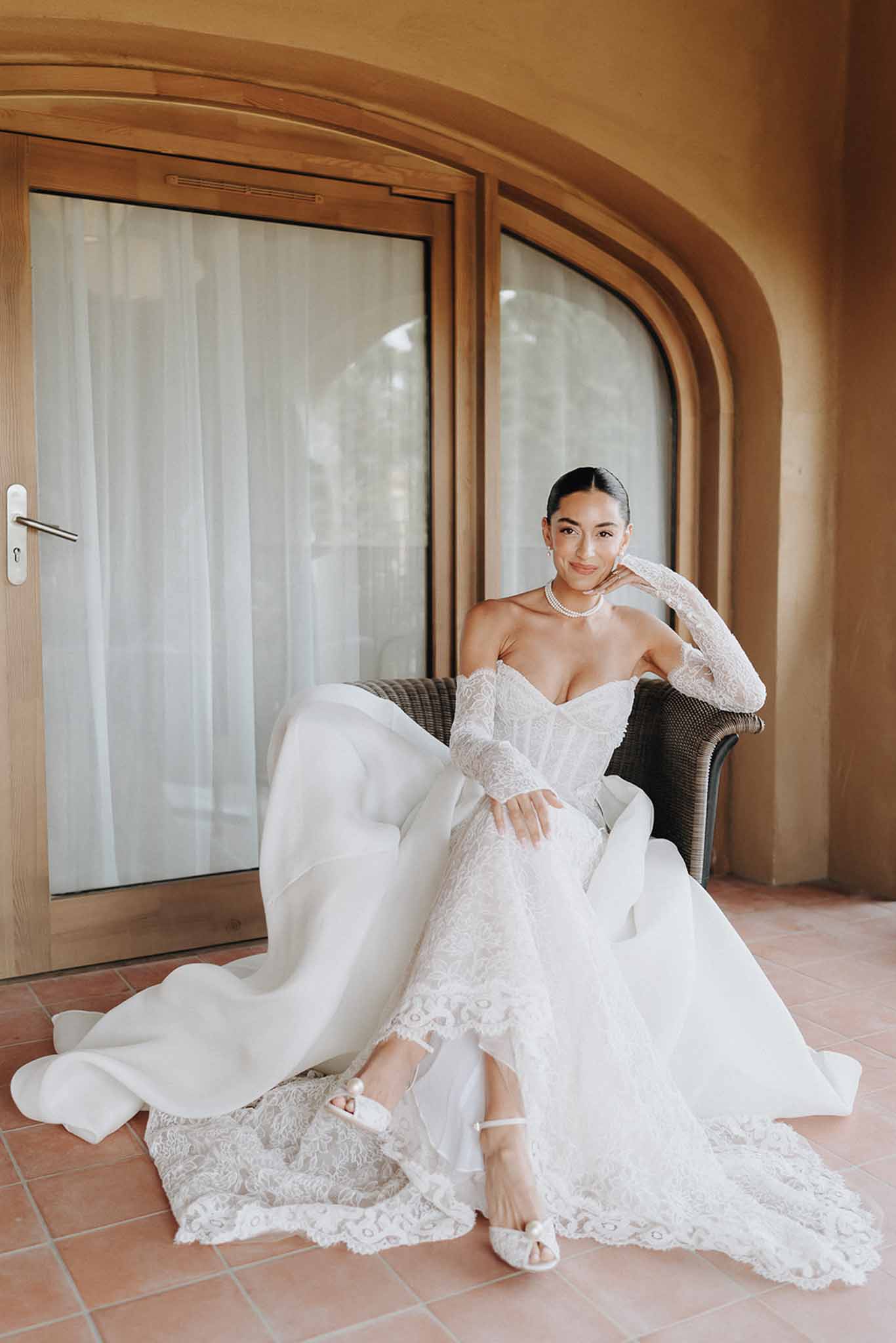 Bride in strapless lace corset gown with detached sleeves and pearl choker seated in wicker armchair