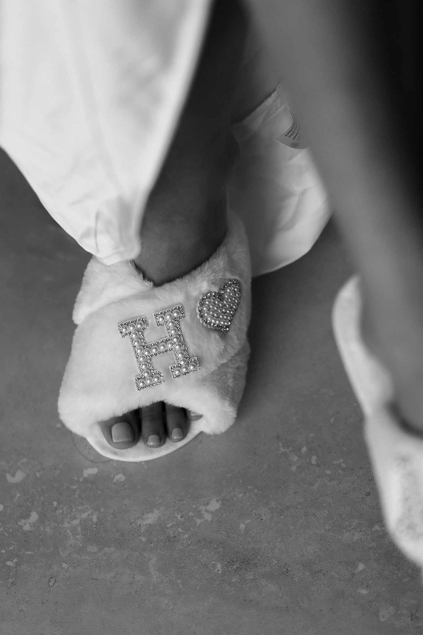 Black-and-white close-up of bride's fluffy open-toe slippers with pearl and rhinestone letter H monogram
