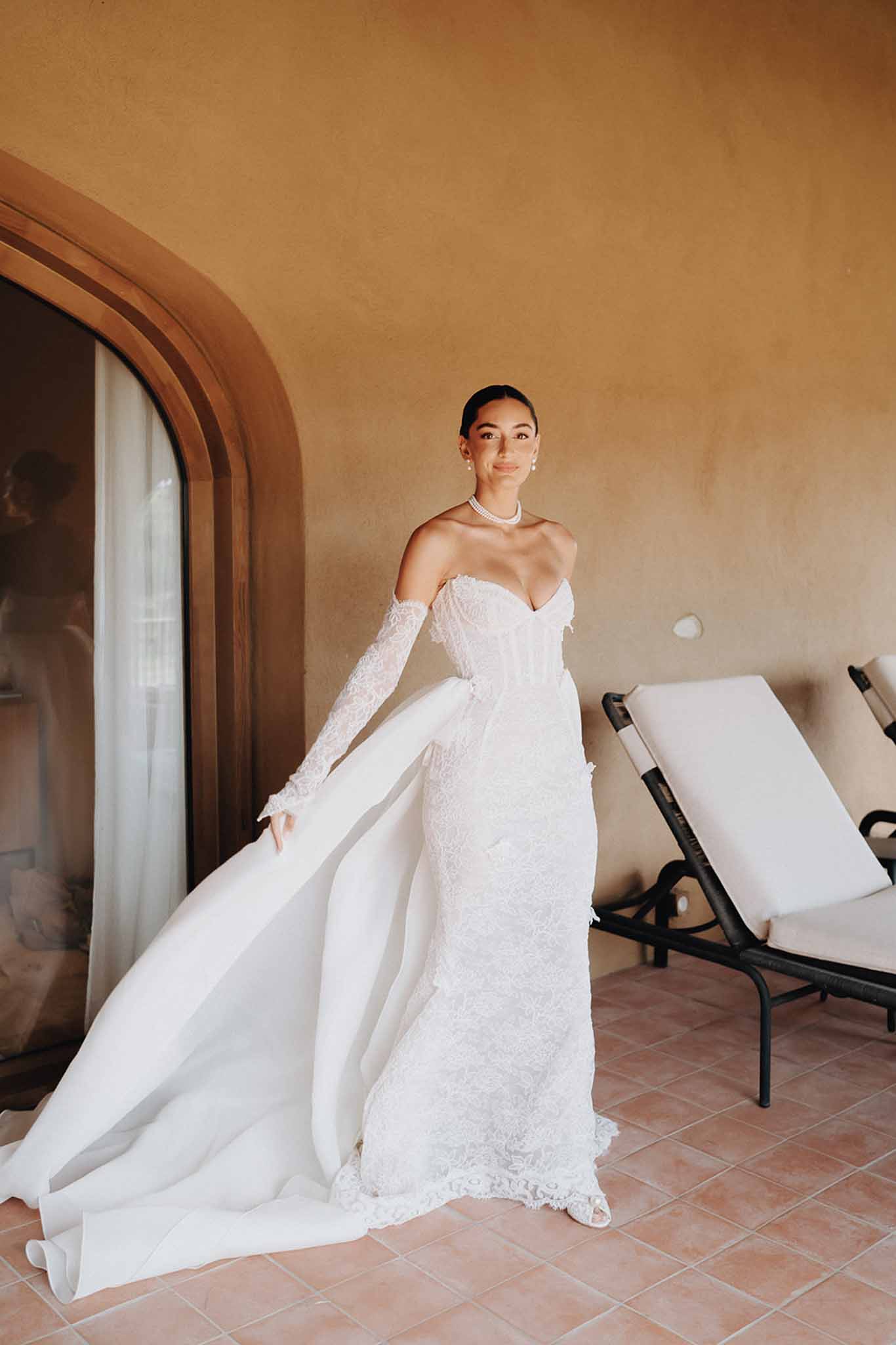 Bride in fitted white lace gown with dramatic overskirt and pearl choker on Mediterranean terrace with ochre walls
