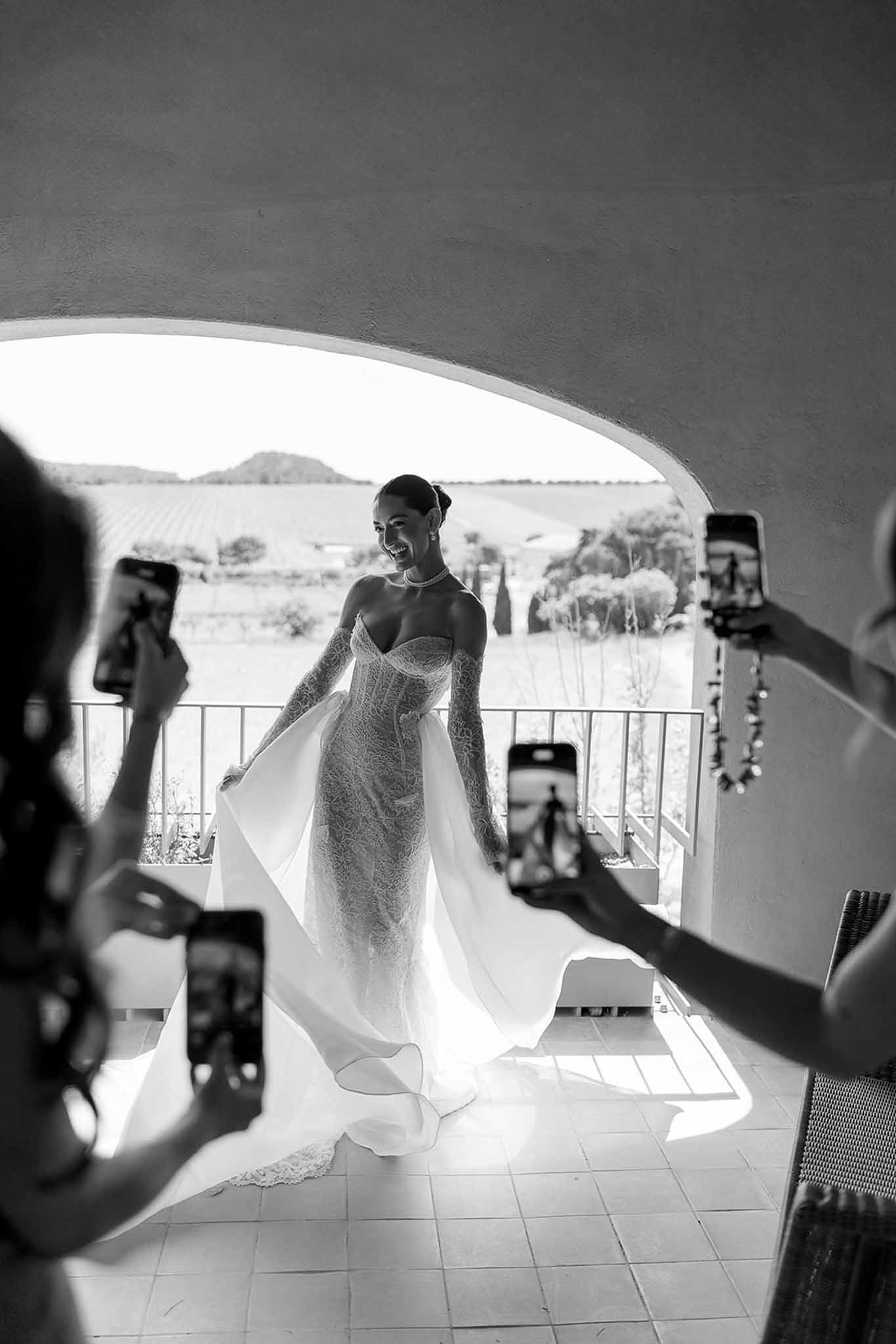 Black and white bride revealing lace gown in archway as bridesmaids photograph her with backlit vineyard view