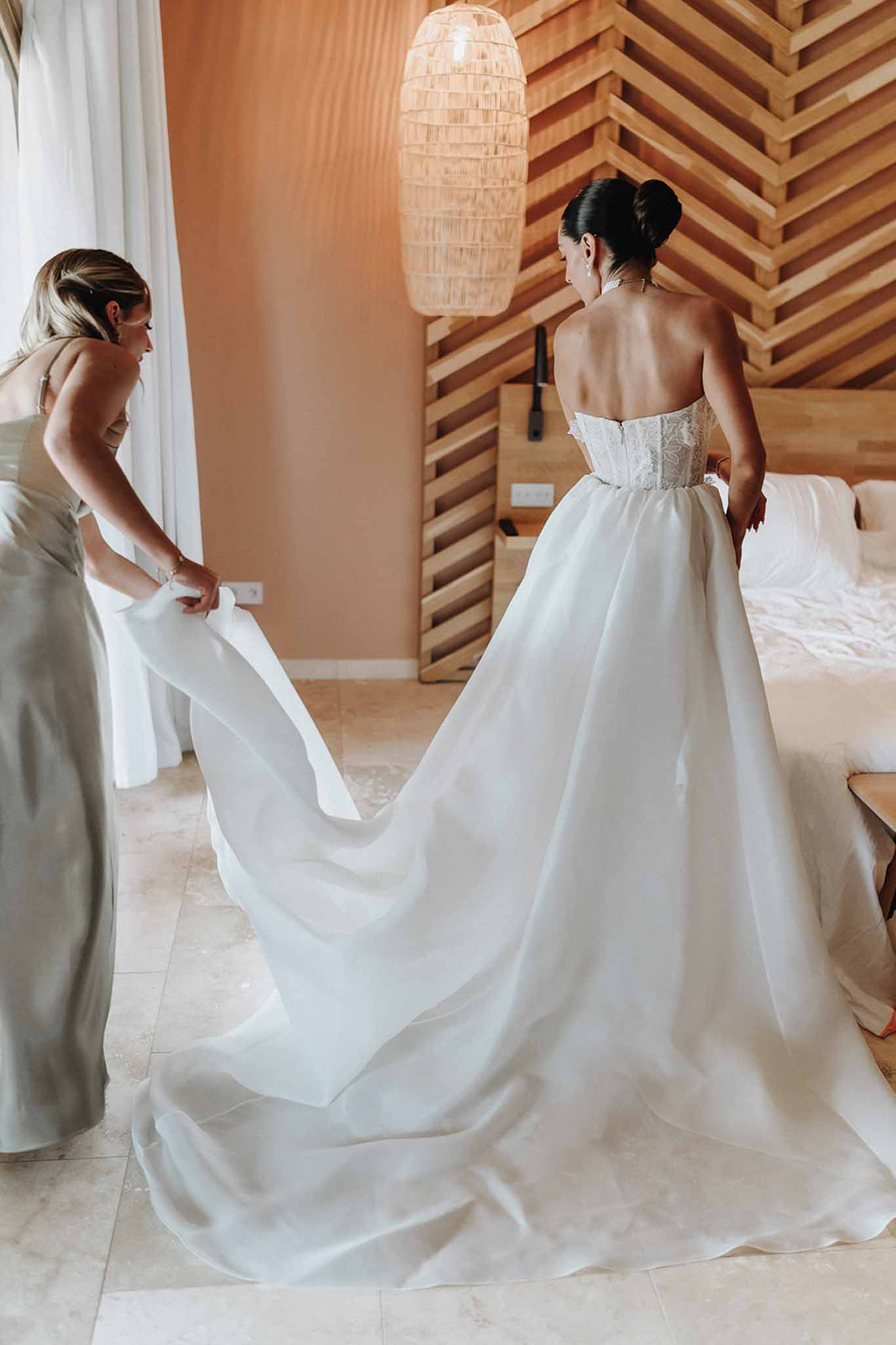 Bridesmaid in sage satin dress arranges train of bride's strapless lace ballgown in modern bedroom