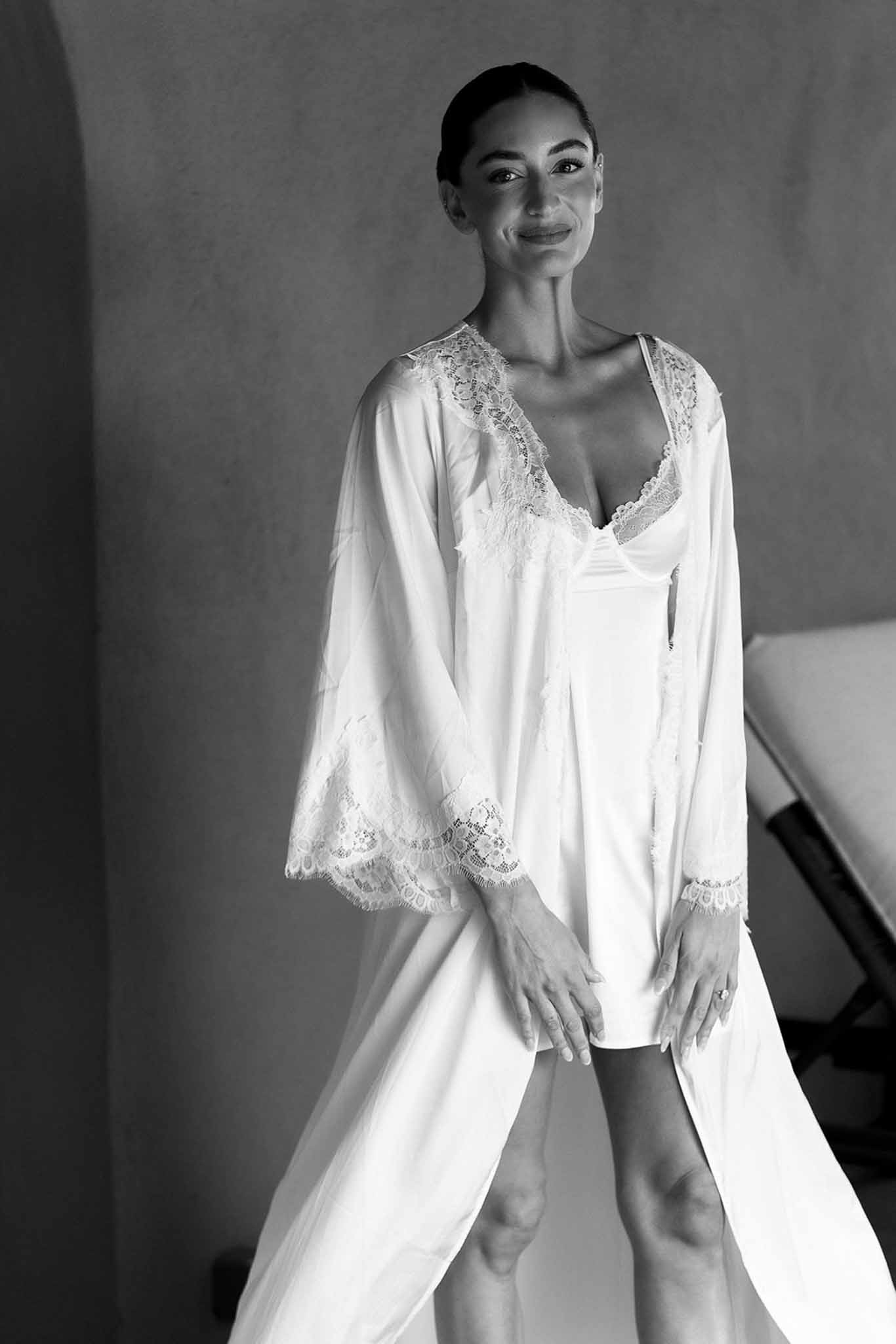Black and white bride in floor-length white satin robe with lace trim standing against plain wall