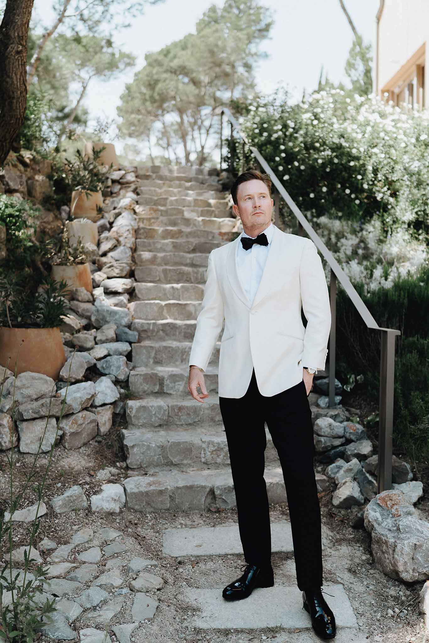 Groom in white dinner jacket and black trousers stands at base of stone garden staircase