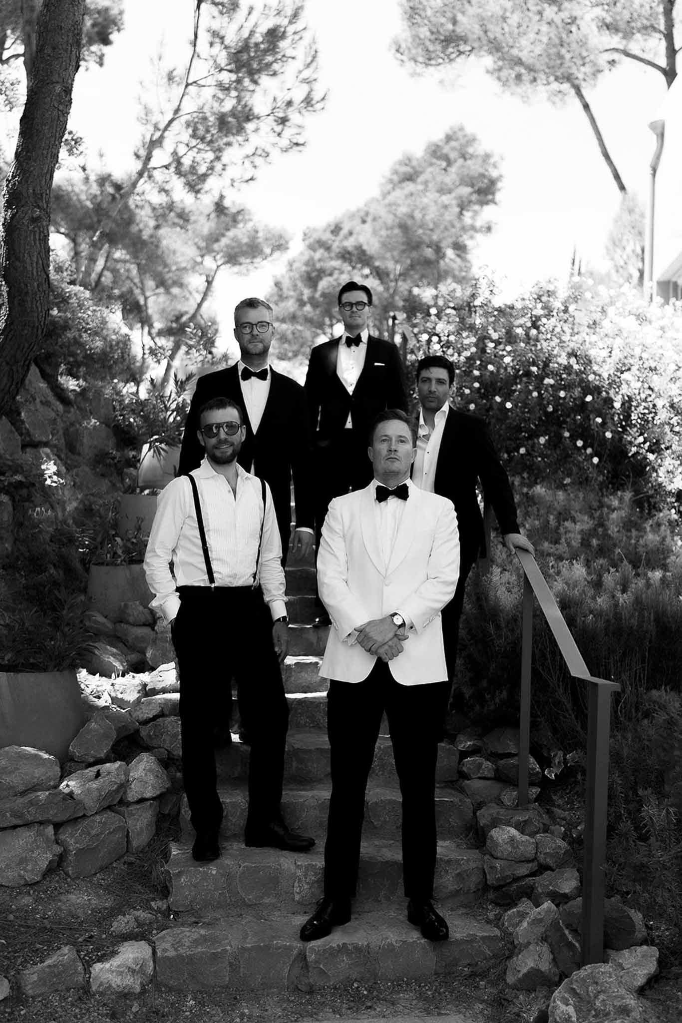 Black and white of groom in white dinner jacket with four groomsmen in tuxedos on garden stone steps