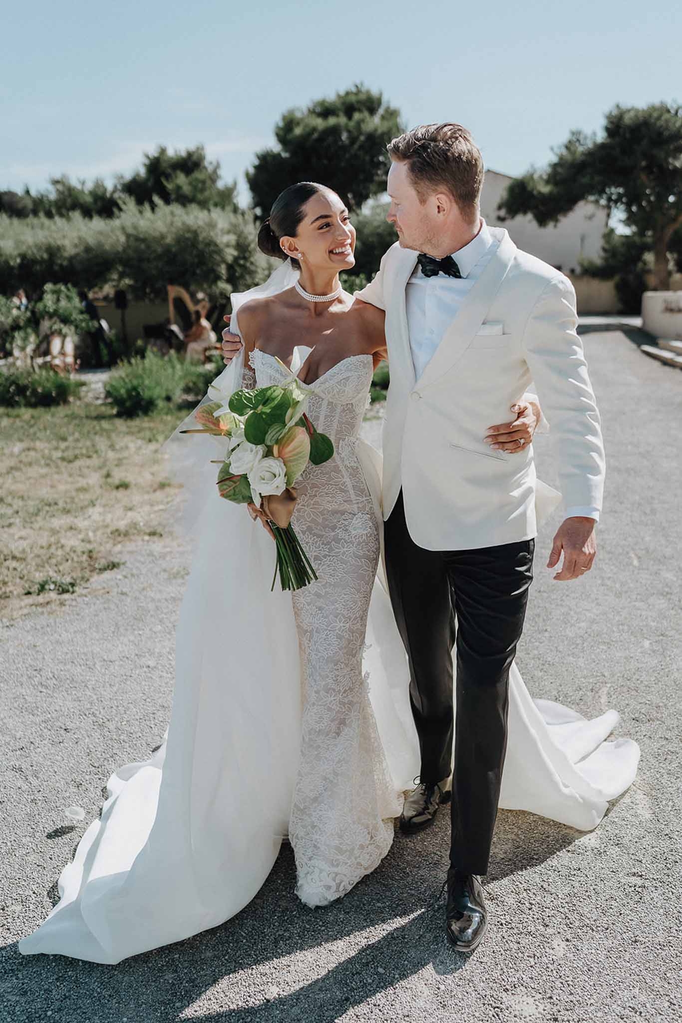 Bride in ivory lace mermaid gown and groom in white dinner jacket walking arm in arm on a gravel path