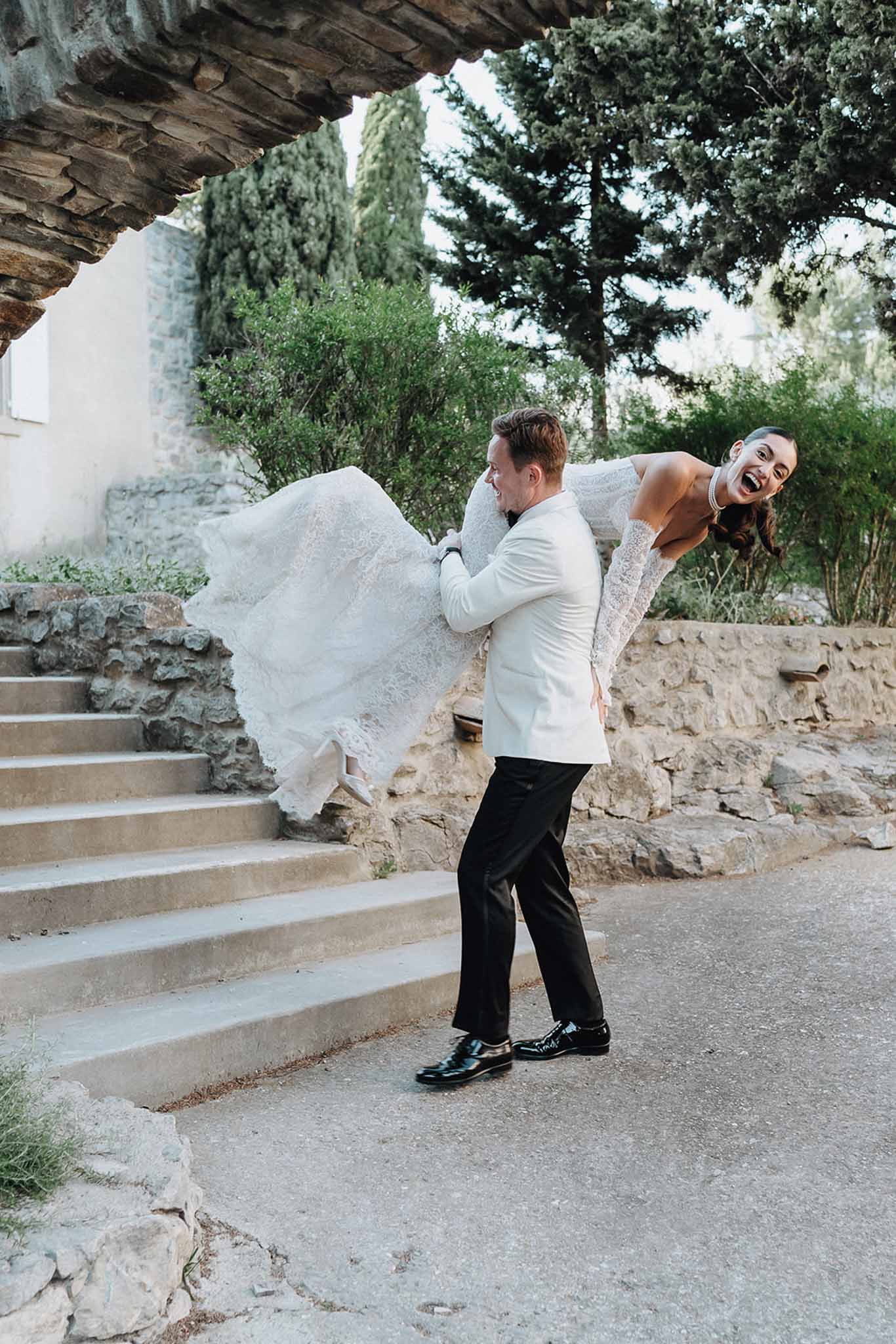 Groom in white dinner jacket lifting laughing bride in lace gown on stone steps at a Provencal property