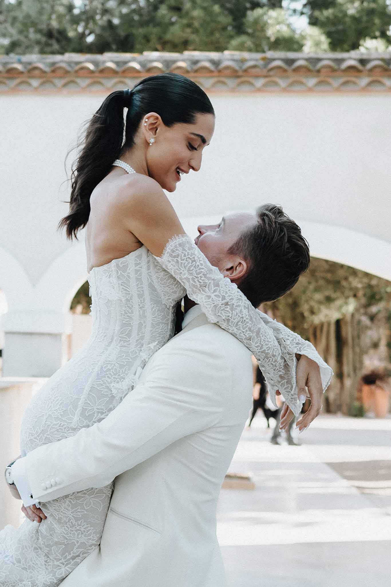 Groom in cream suit lifting laughing bride in off-shoulder lace gown with pearl choker at Provencal venue