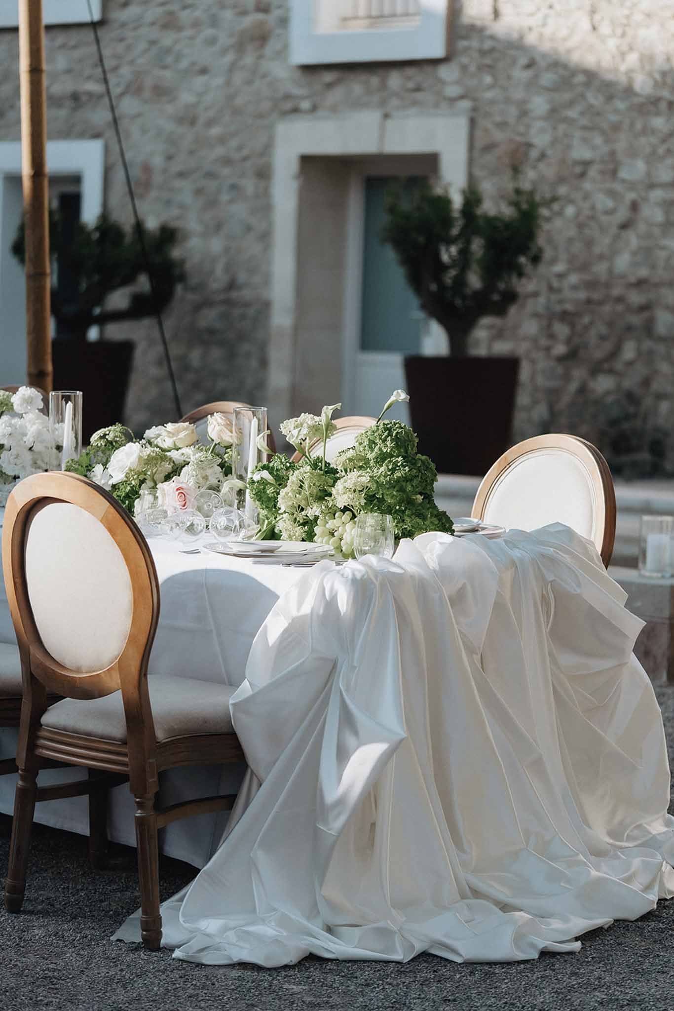Outdoor reception head table with white linen, green hydrangeas, white calla lilies, ranunculus, and grapes centerpiece