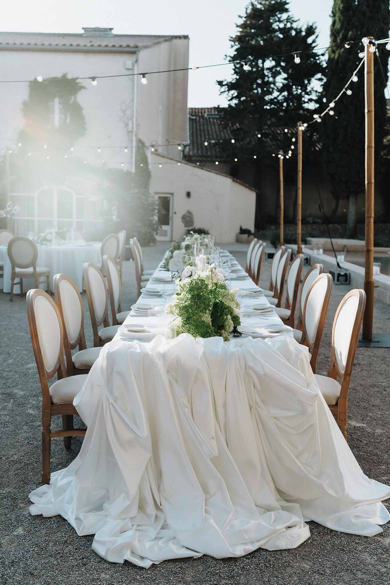 Long feasting table with draped ivory cloth and green hydrangea centerpieces under string lights in Provencal courtyard