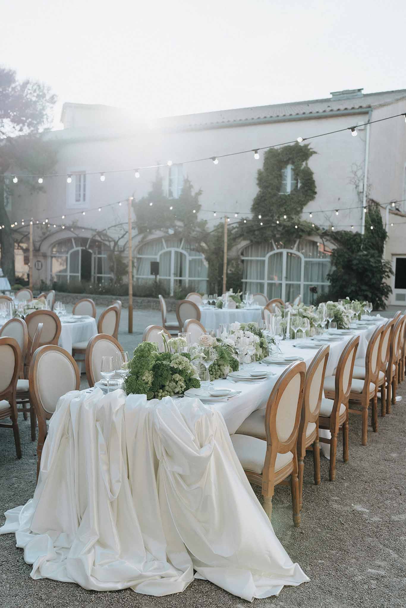 Long silk-draped table with green hydrangea runner and medallion chairs under fairy lights at dusk