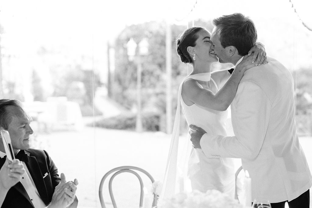 Black-and-white candid of bride and groom kissing during reception speech with guest applauding