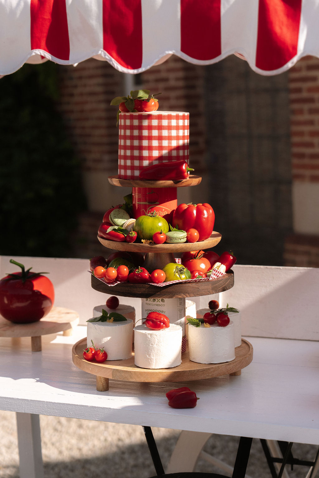 Italian-themed wedding dessert display with tiered wooden stand, tomatoes, macarons, and cheese under striped awning