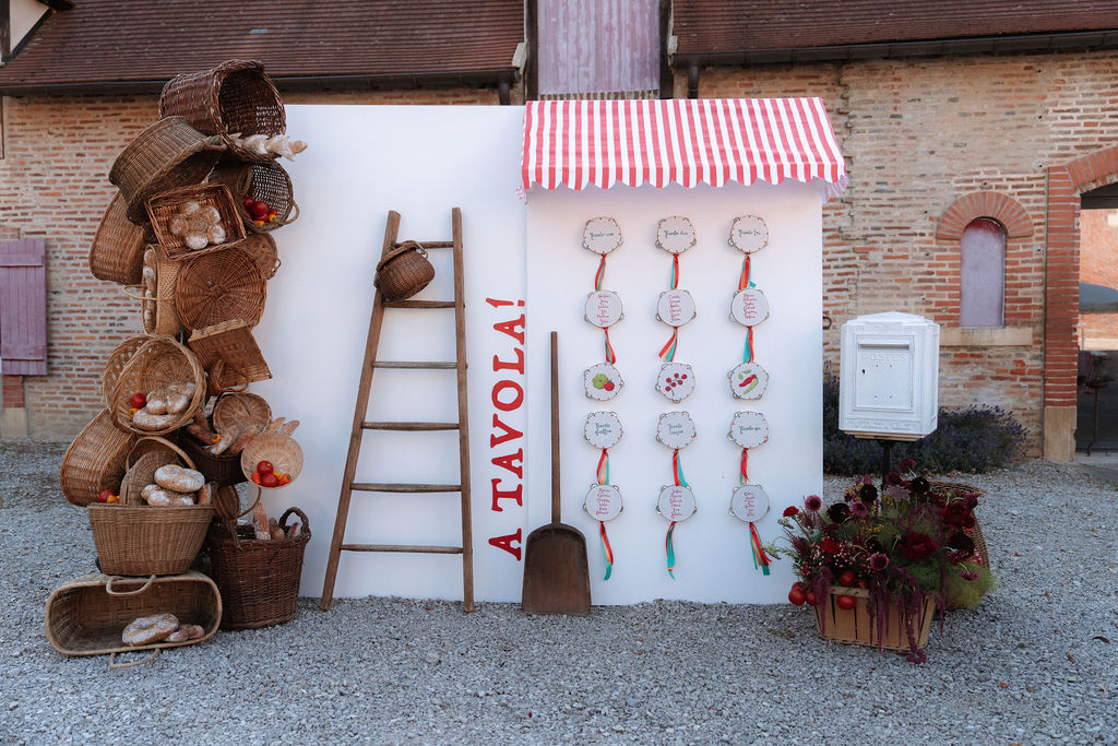 Italian-themed wedding seating chart with A Tavola sign, striped awning, bread baskets, and burgundy florals