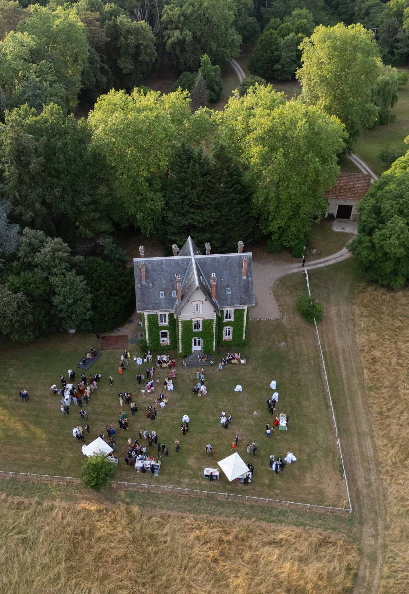 Aerial golden hour view of 100 guests at cocktail hour on lawn of ivy-covered French manor
