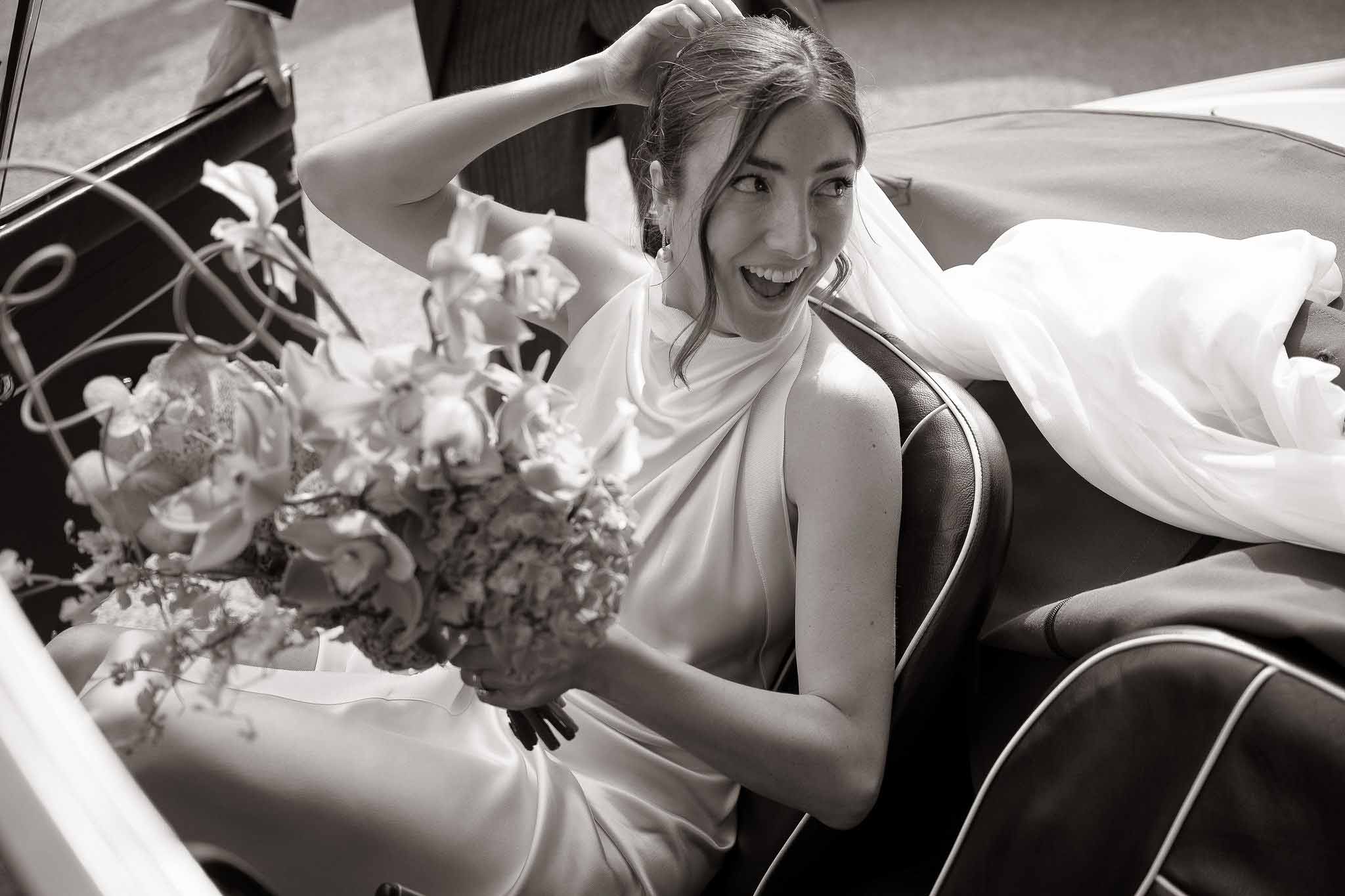 Black and white portrait of laughing bride holding bouquet inside vintage car with flowing veil