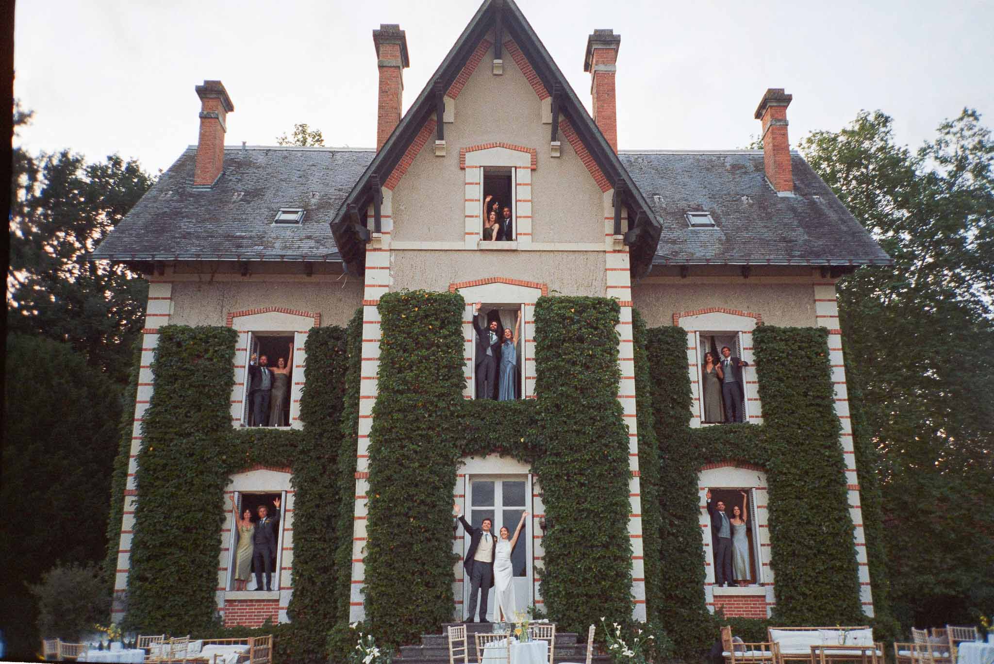 Couple celebrating at entrance while bridal party waves from windows across three-storey manor facade