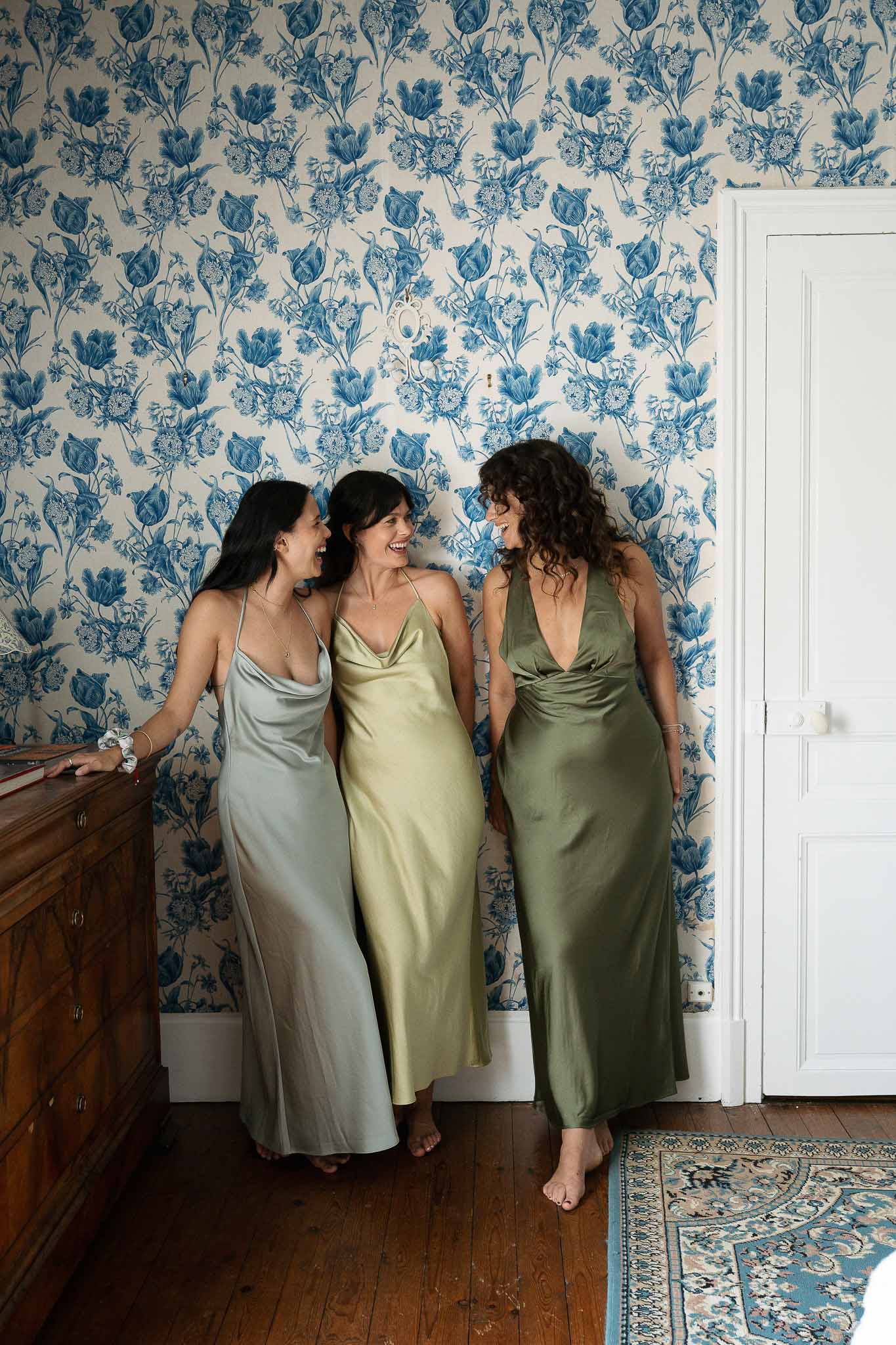Three bridesmaids in tonal green satin slip dresses laughing in blue toile wallpaper chateau bedroom