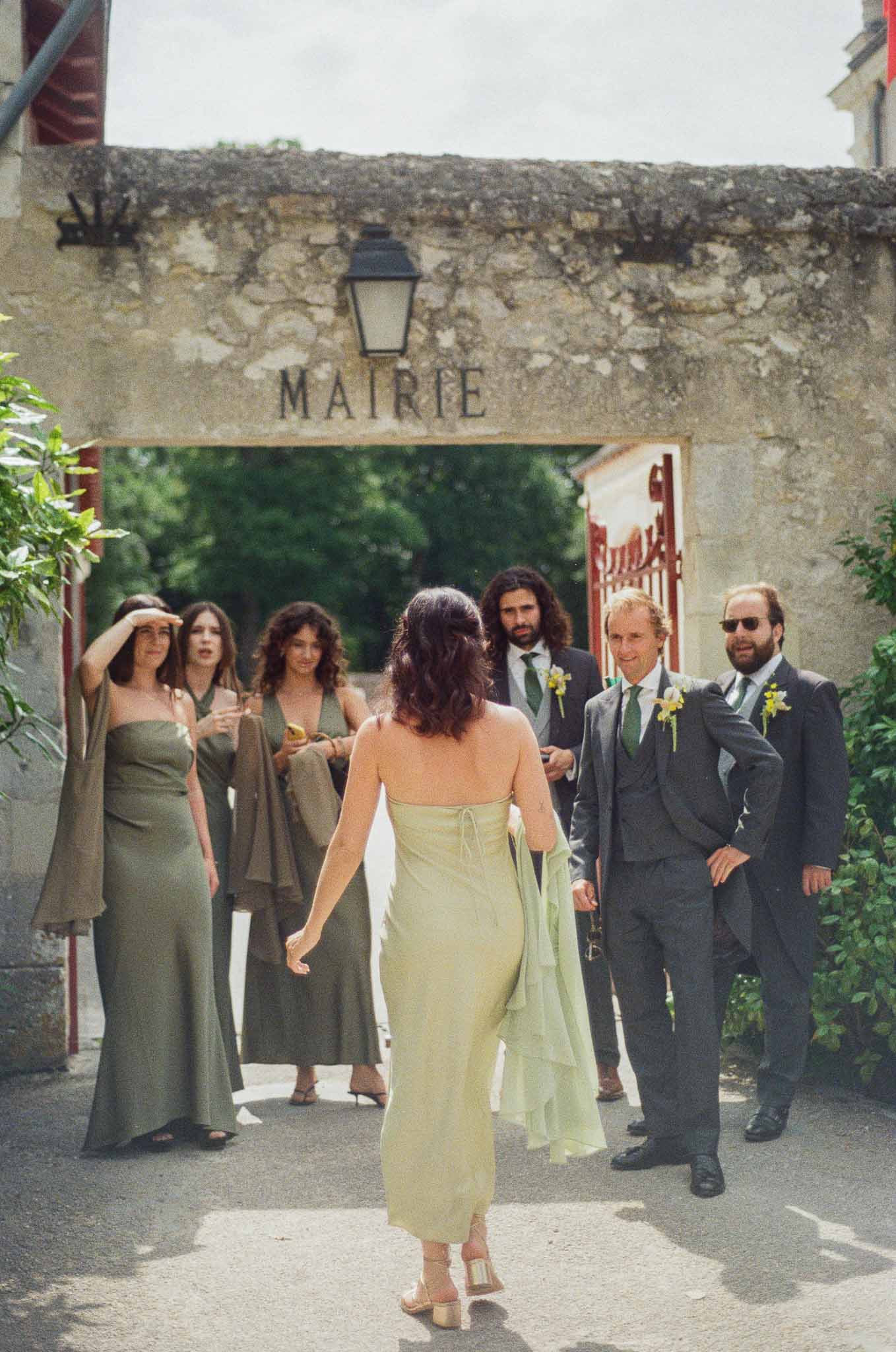 Three bridesmaids in olive and sage dresses with three groomsmen in charcoal suits outside stone mairie entrance