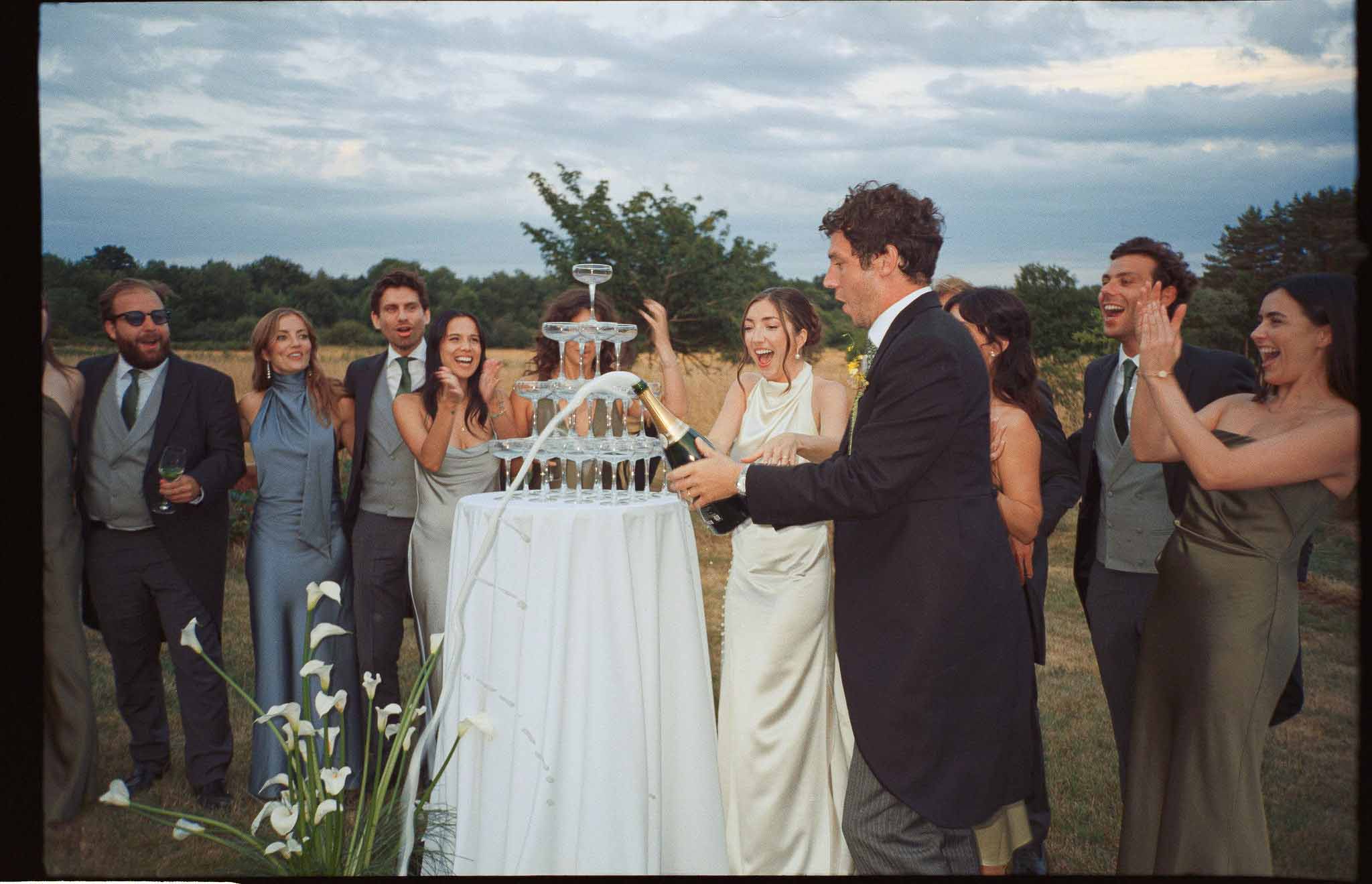 Bride and groom pouring champagne into a coupe glass tower at outdoor cocktail hour while guests cheer and clap