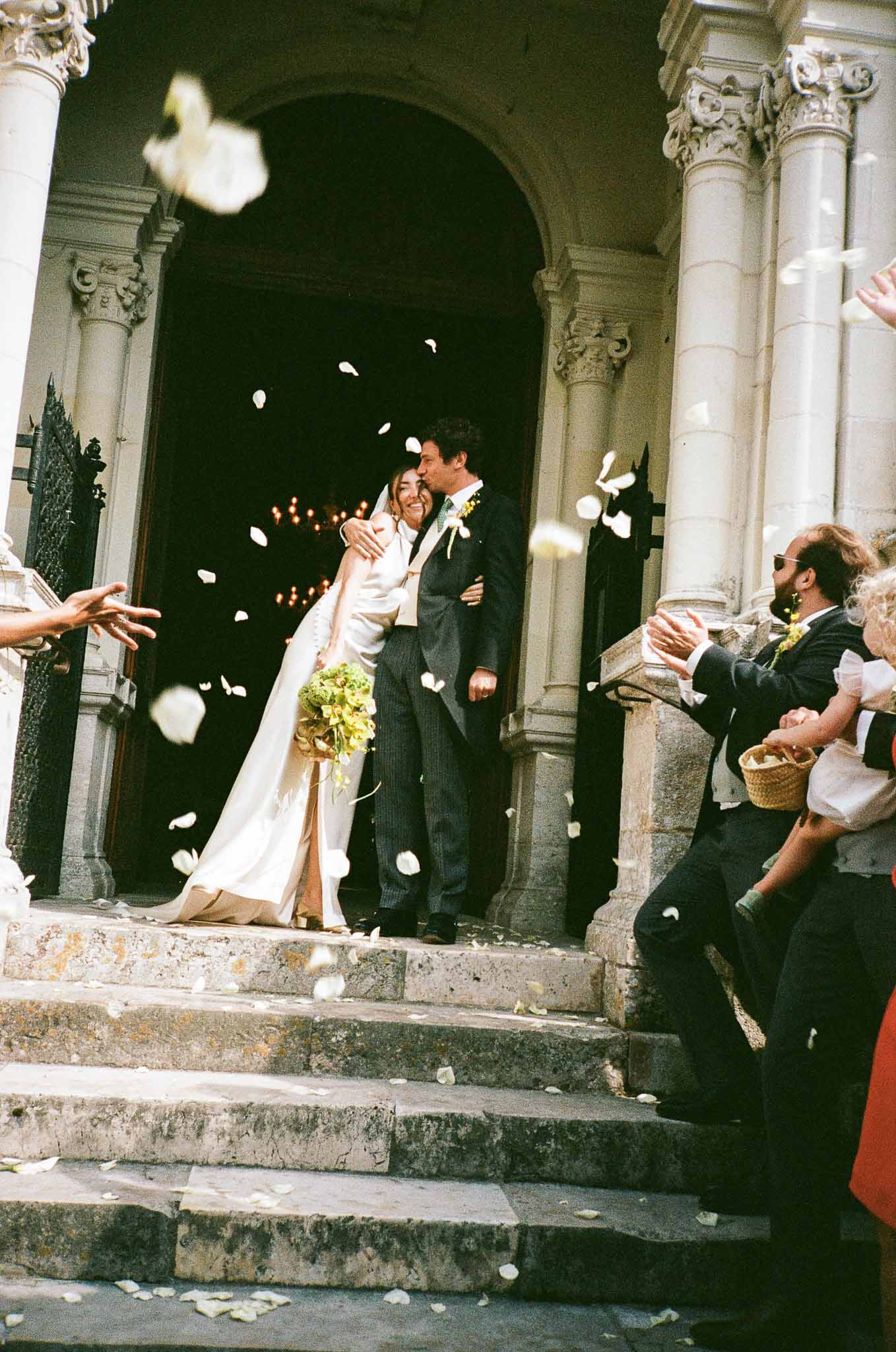 Couple exits stone church doorway as guests throw white rose petals, bride carrying chartreuse orchid bouquet