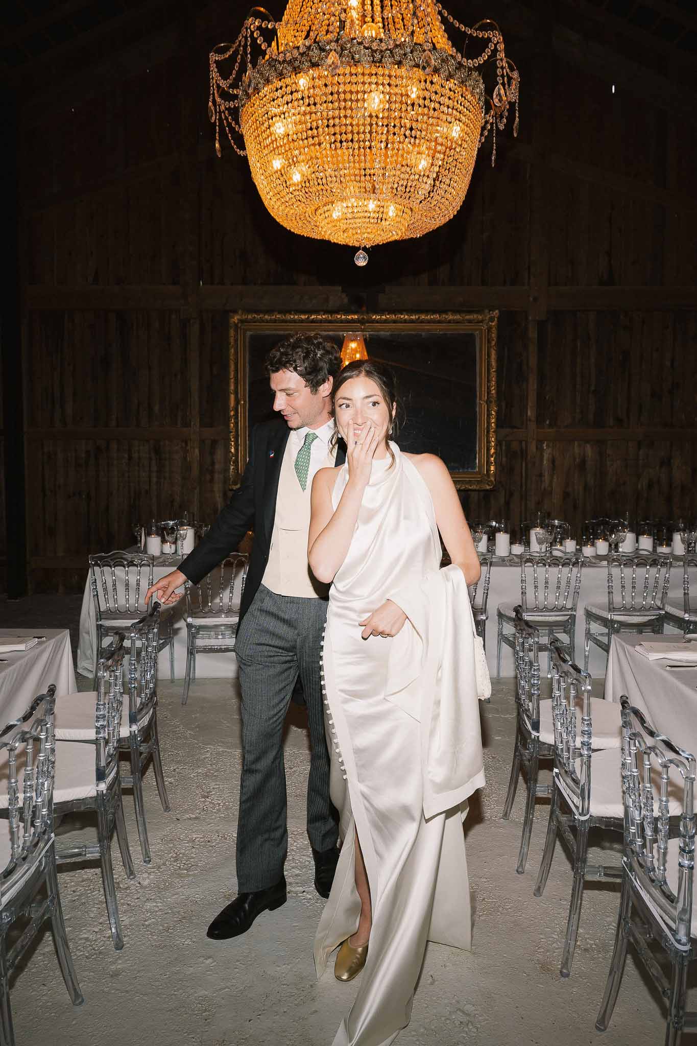 Bride and groom see decorated barn reception for first time with gold chandelier and acrylic chairs