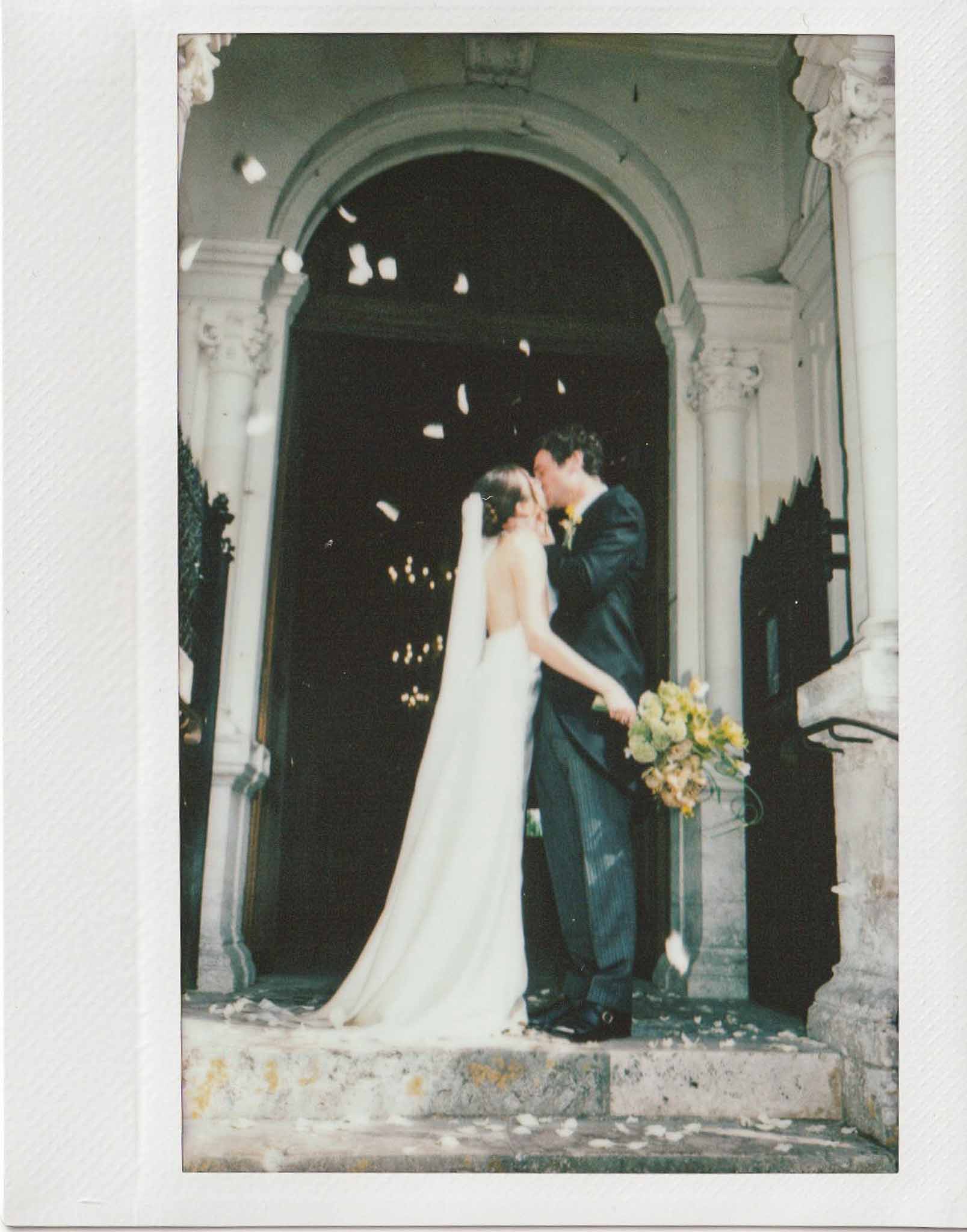 Polaroid-style photo of bride and groom kissing on stone steps with confetti petals and cathedral veil