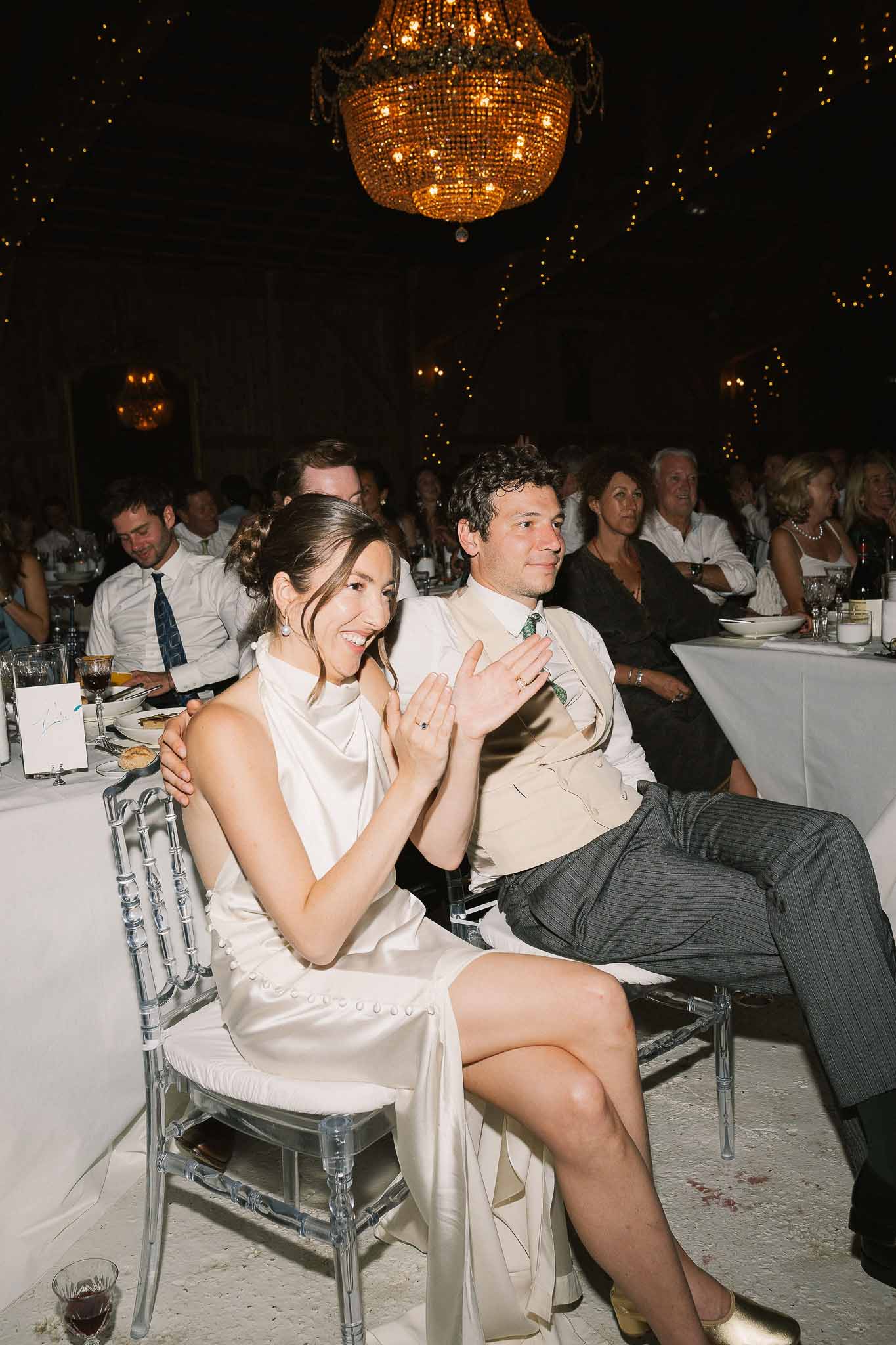 Bride and groom clapping during speeches beneath amber chandelier and fairy lights at indoor reception