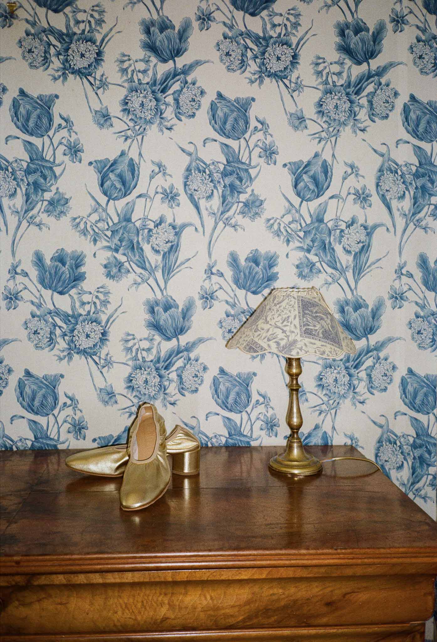 Gold block-heel bridal shoes beside brass lamp on walnut dresser with blue toile de Jouy wallpaper
