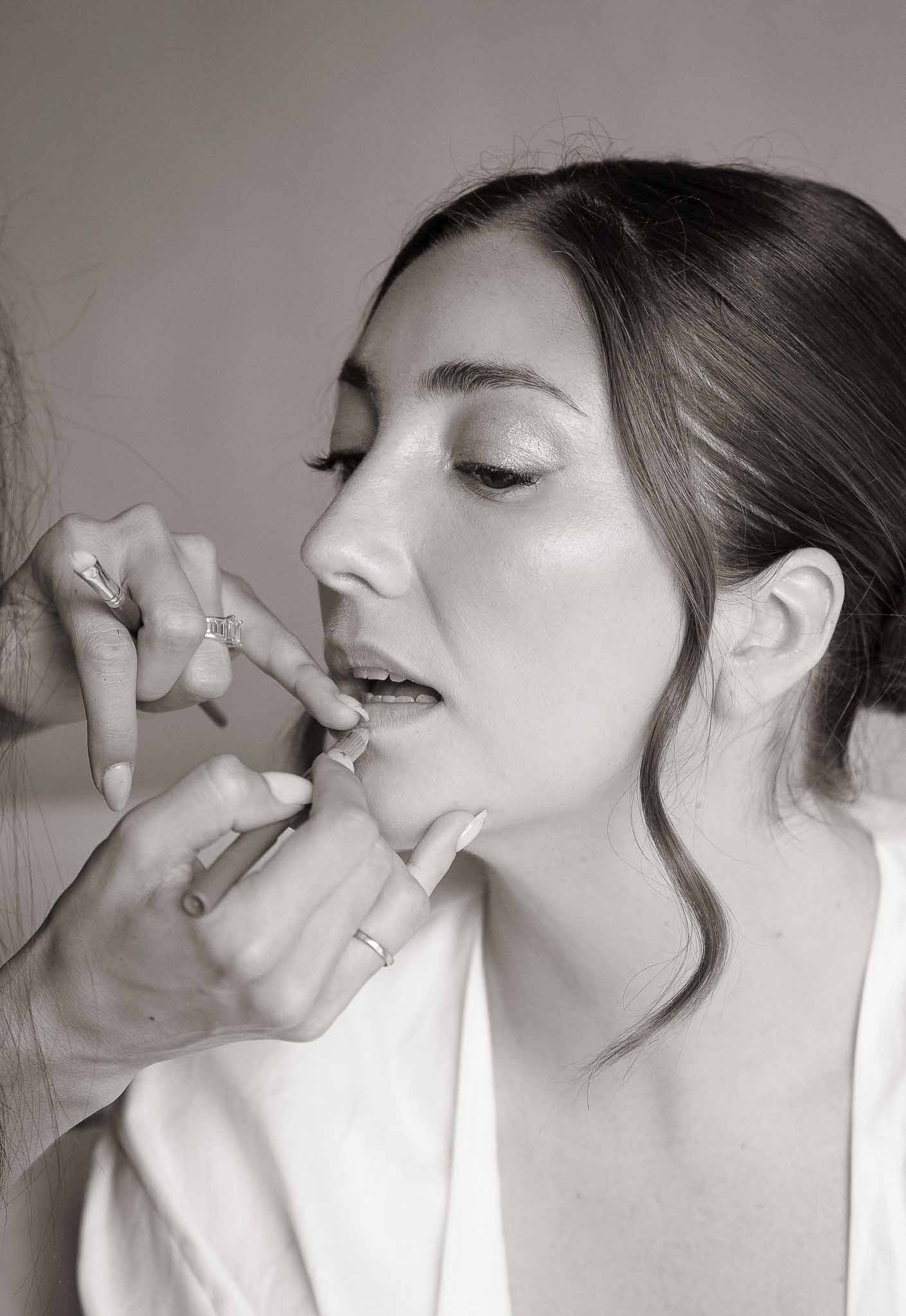 Black-and-white close-up of makeup artist applying lipstick to bride wearing white robe during getting ready