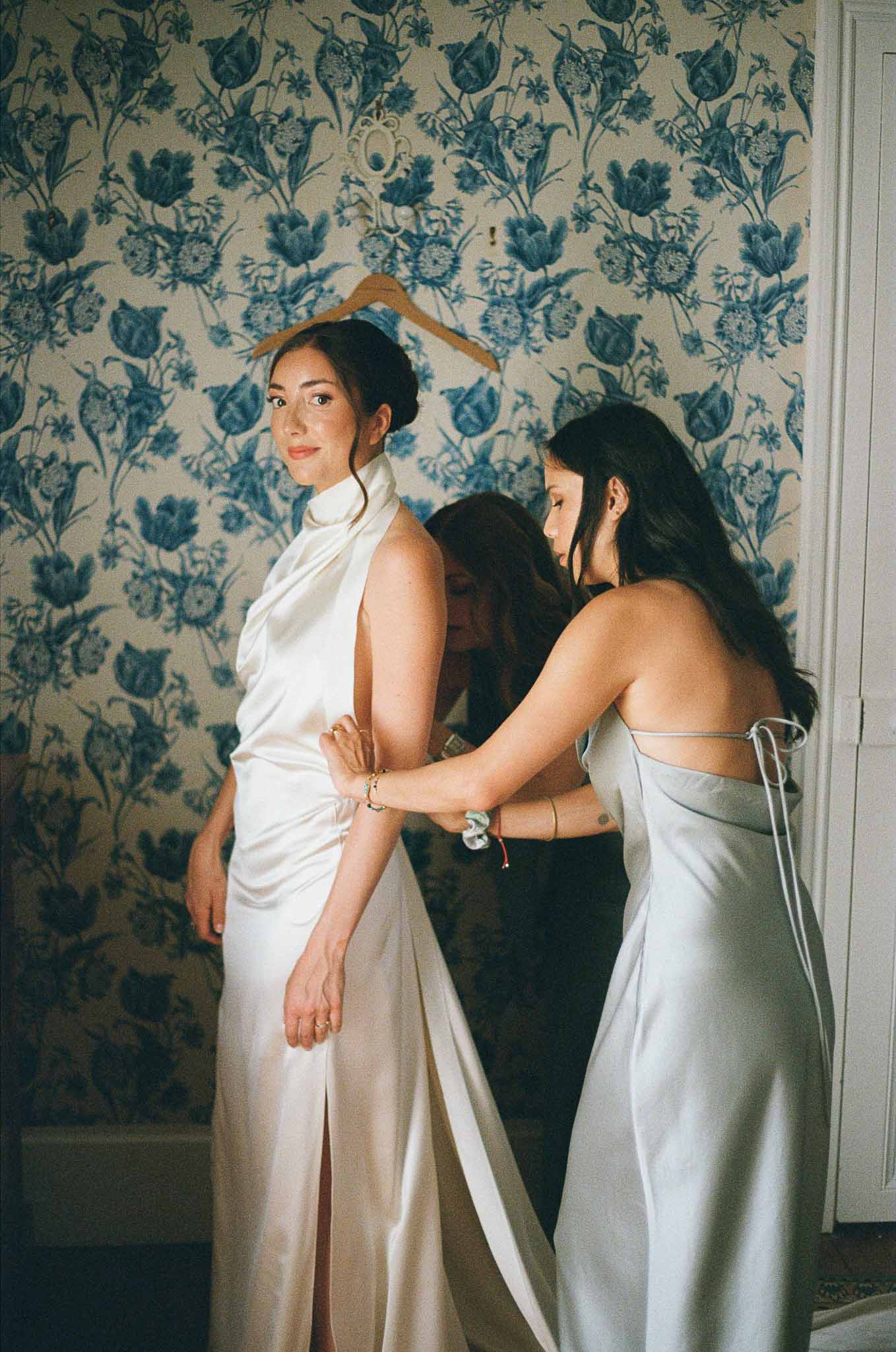 Two bridesmaids fasten bride's satin halter gown in toile-wallpapered chateau room in warm film tones