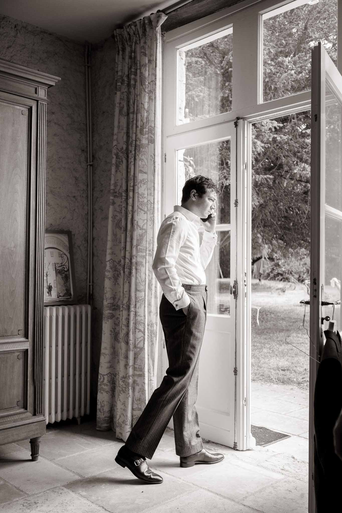 Black and white of groom in shirt and cufflinks on phone at French door in toile-curtained chateau room