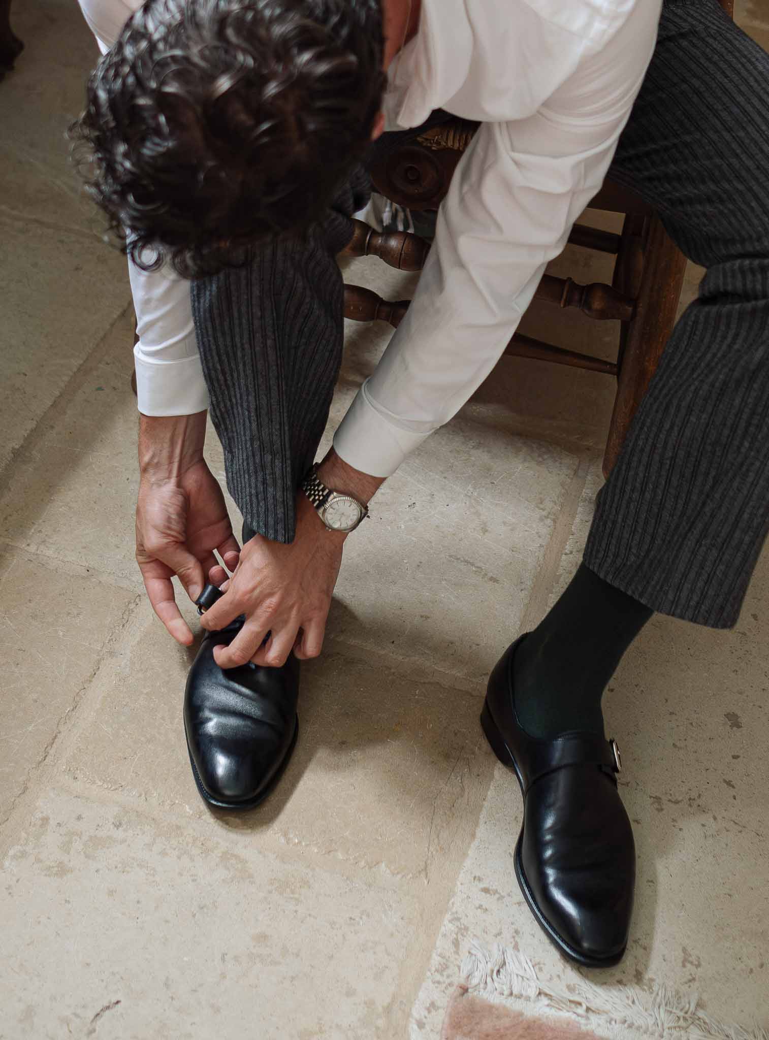 Groom fastening black leather monk strap shoes wearing charcoal pinstripe trousers and white dress shirt
