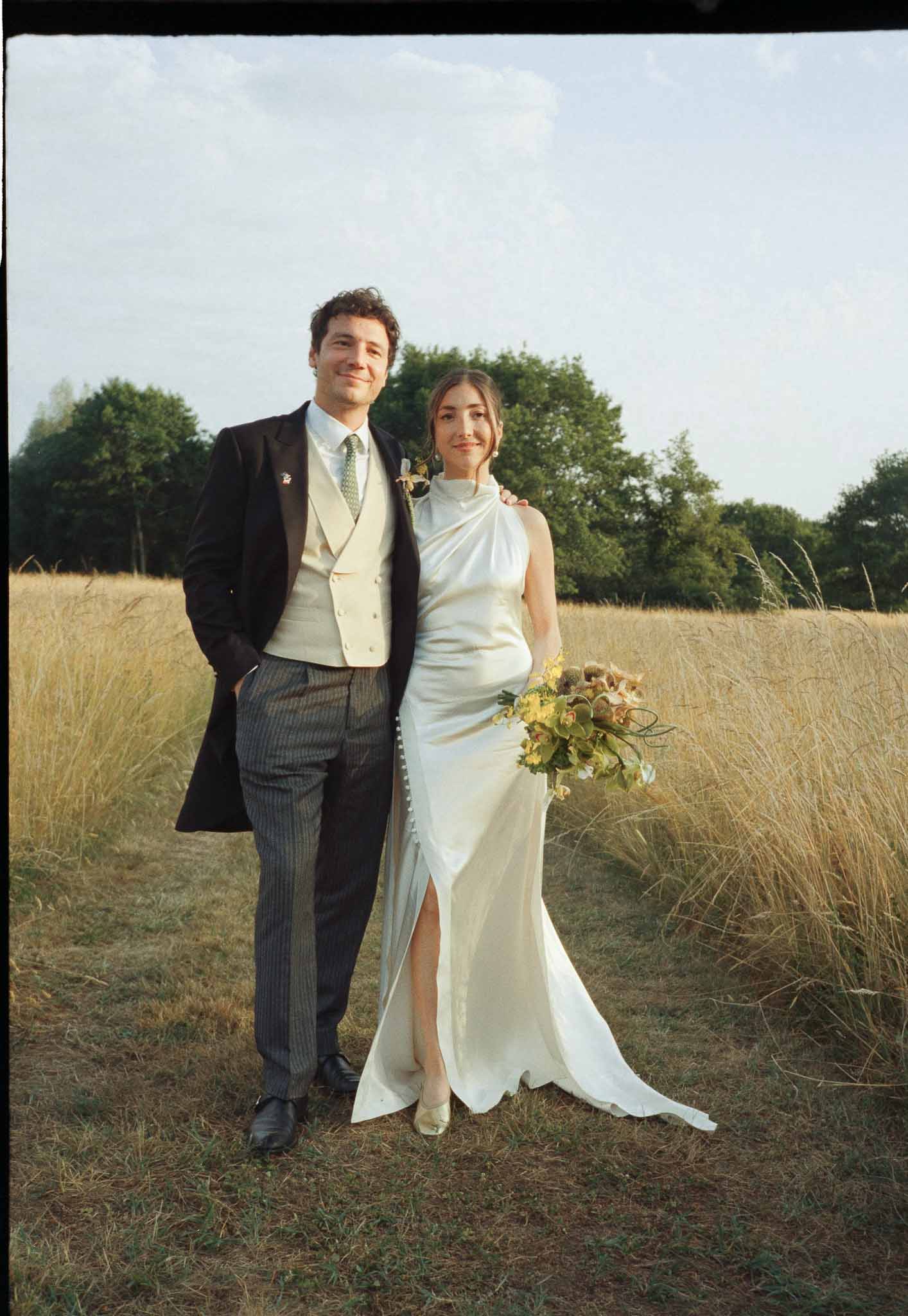 Couple in golden grass field bride in ivory halter satin gown holding orchid bouquet groom in black morning coat