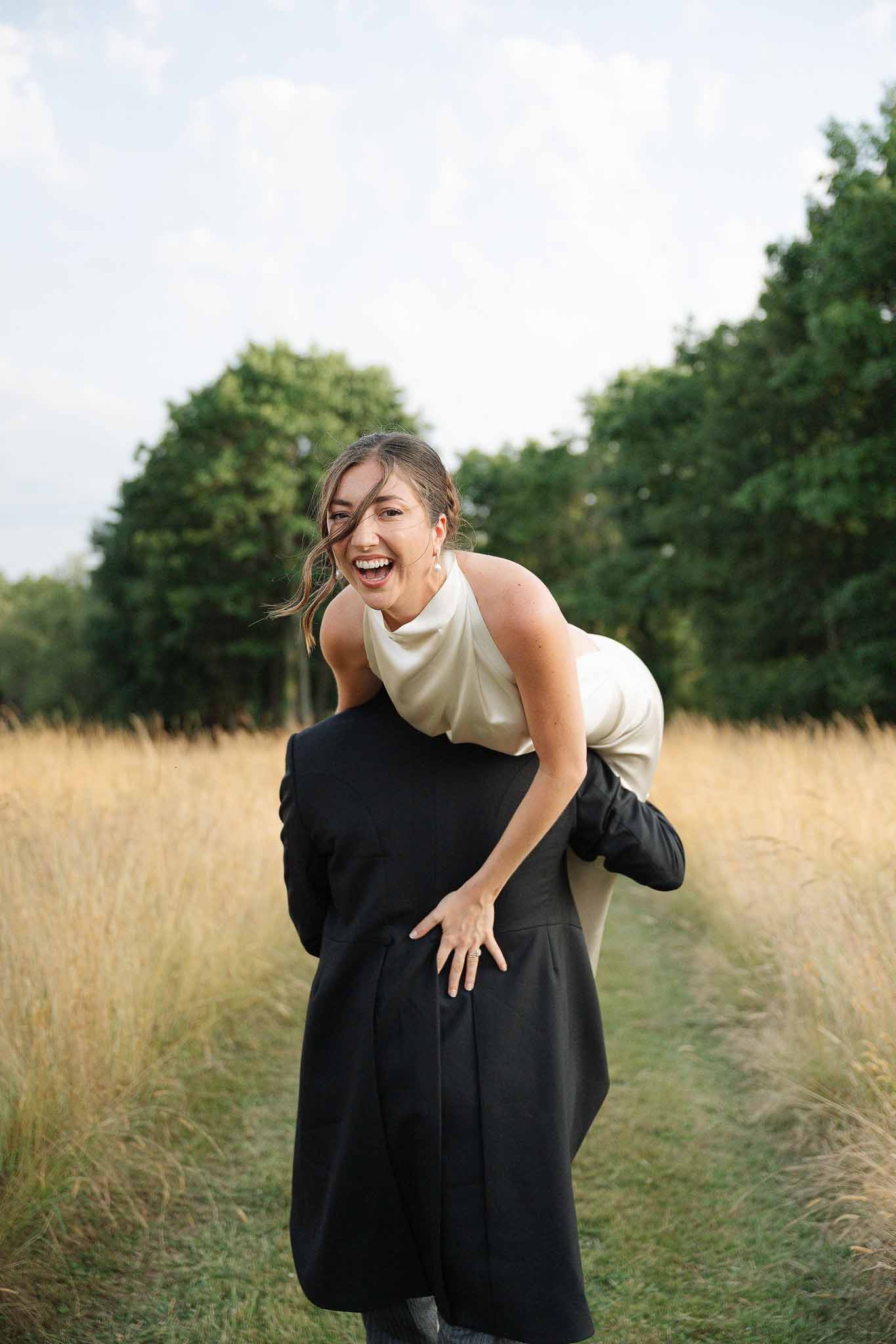 Groom carrying bride over shoulder in golden field, bride in ivory halter-neck satin dress laughing