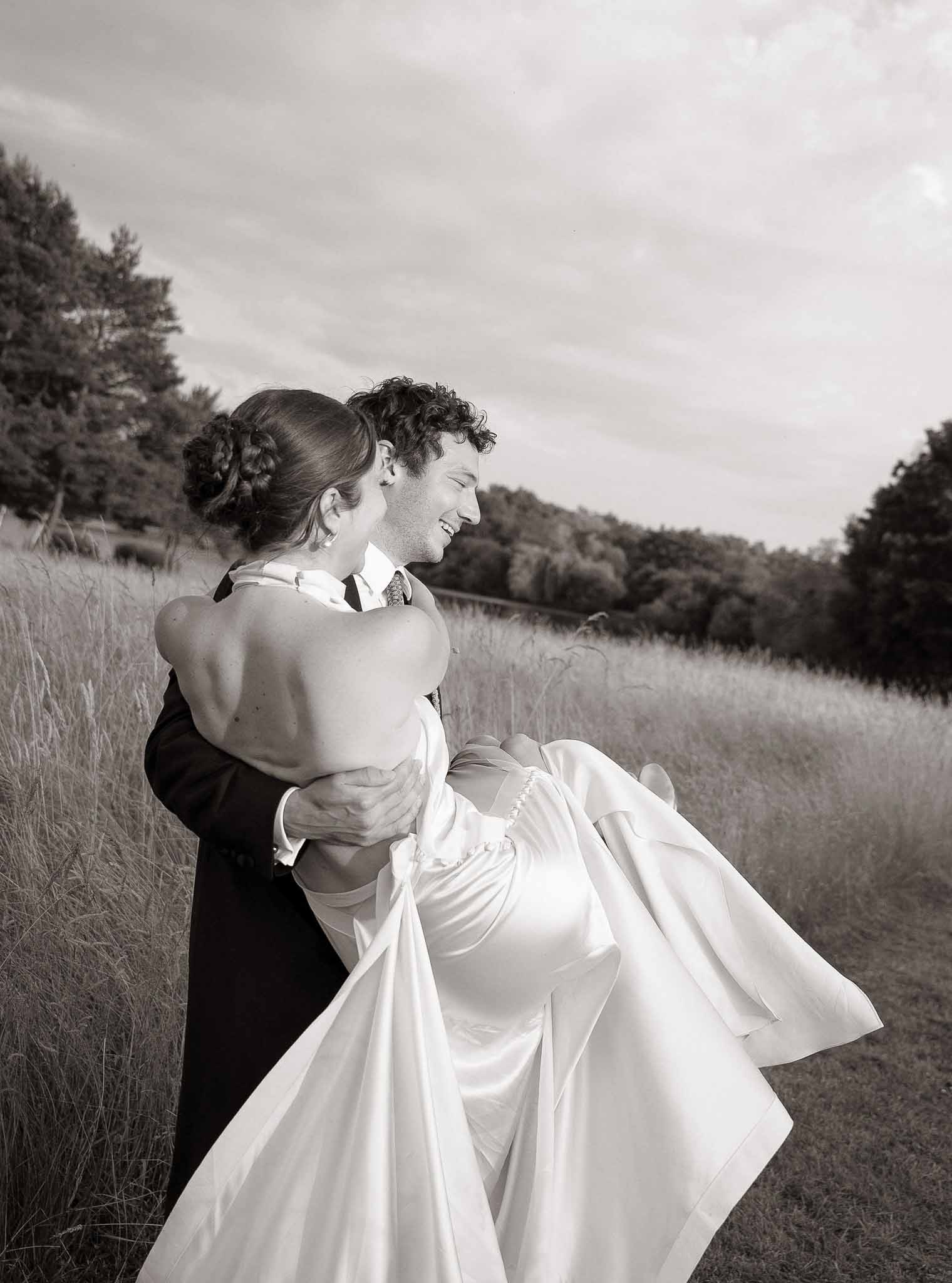 Groom lifts laughing bride in satin halter gown with braided updo in open field in B&W