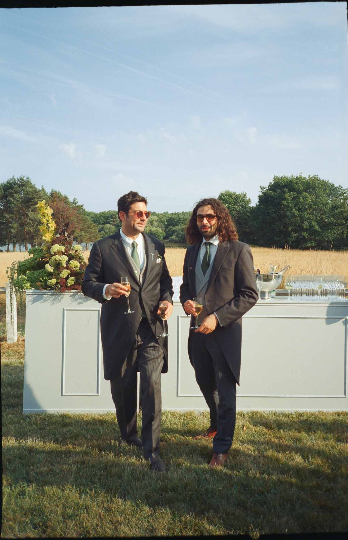 Two male guests in charcoal suits with green ties holding champagne at outdoor bar on film