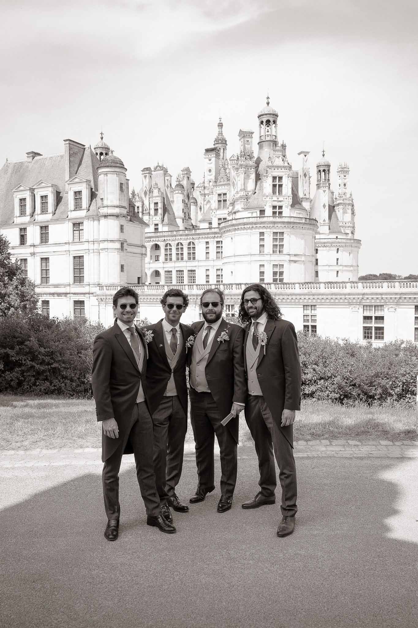 Black and white of four groomsmen in dark suits with sunglasses posing before Chateau de Chambord Renaissance facade