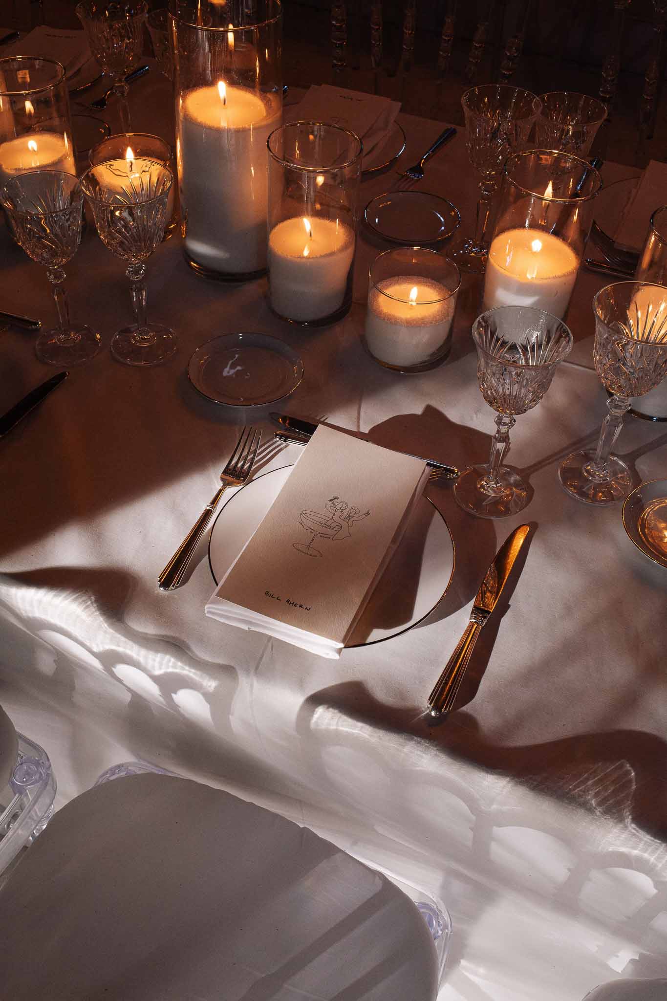 Wedding reception table setting with white pillar candles in hurricane holders, crystal glasses, gold cutlery, and persona...