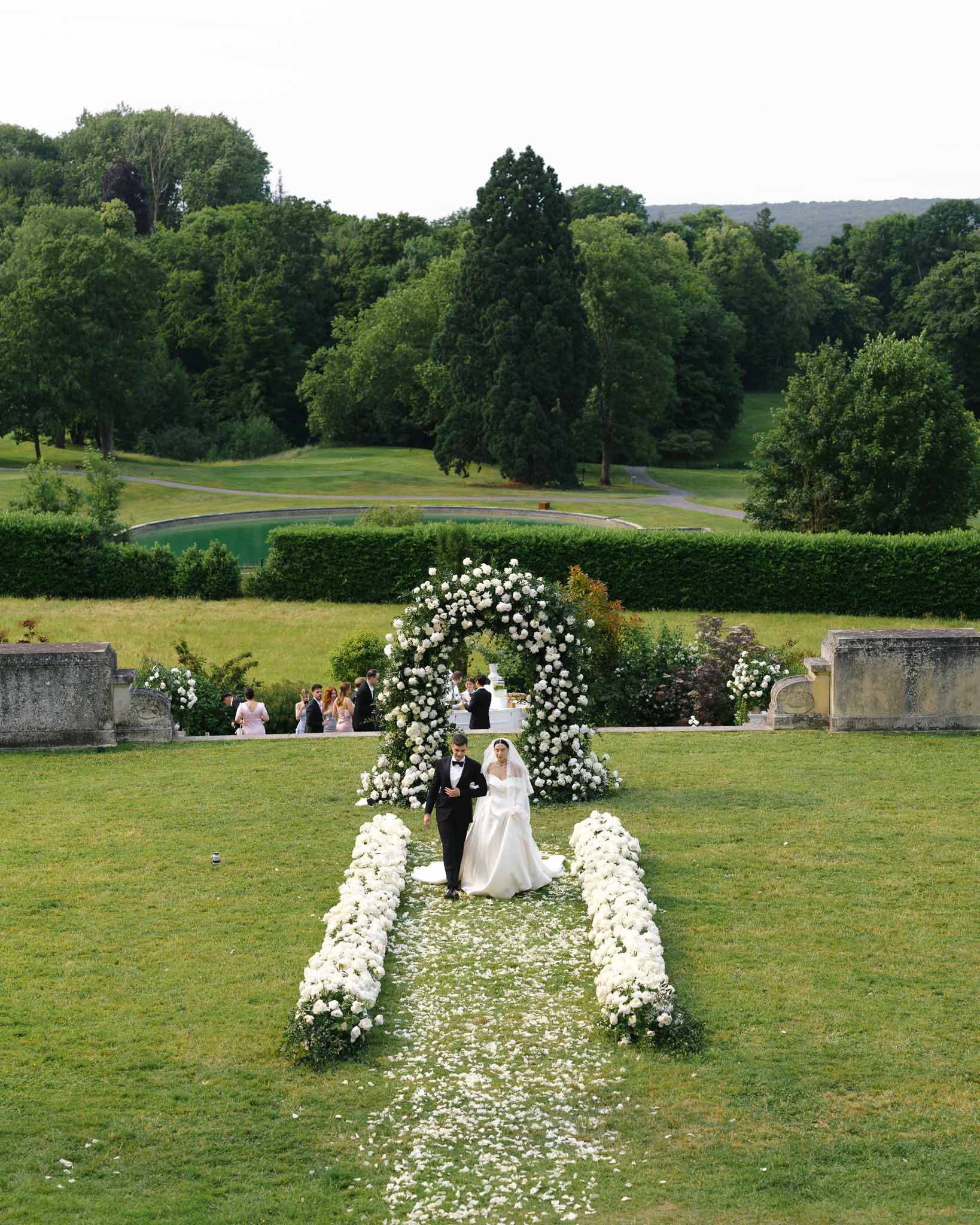 Aerial view of bride and groom walking down a white rose-lined aisle beneath a floral arch on chateau grounds