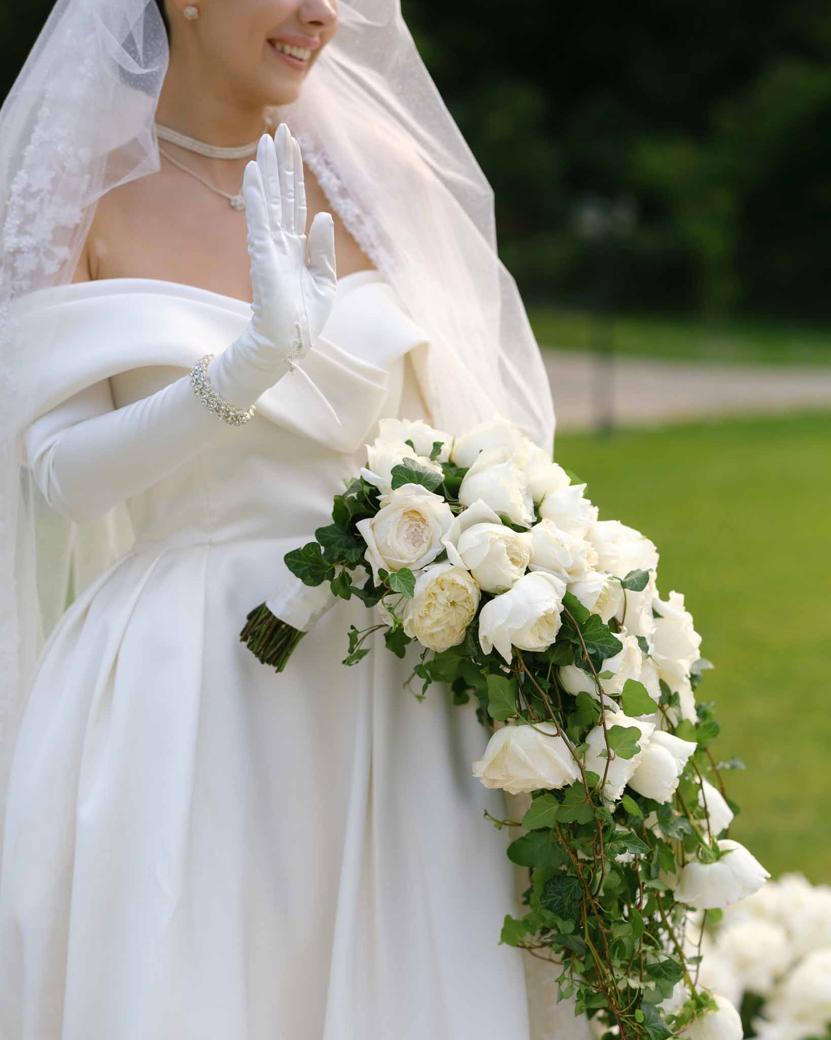 Close-up bride in ivory satin gown with bow detail, satin gloves, and cascading white rose and ivy bouquet