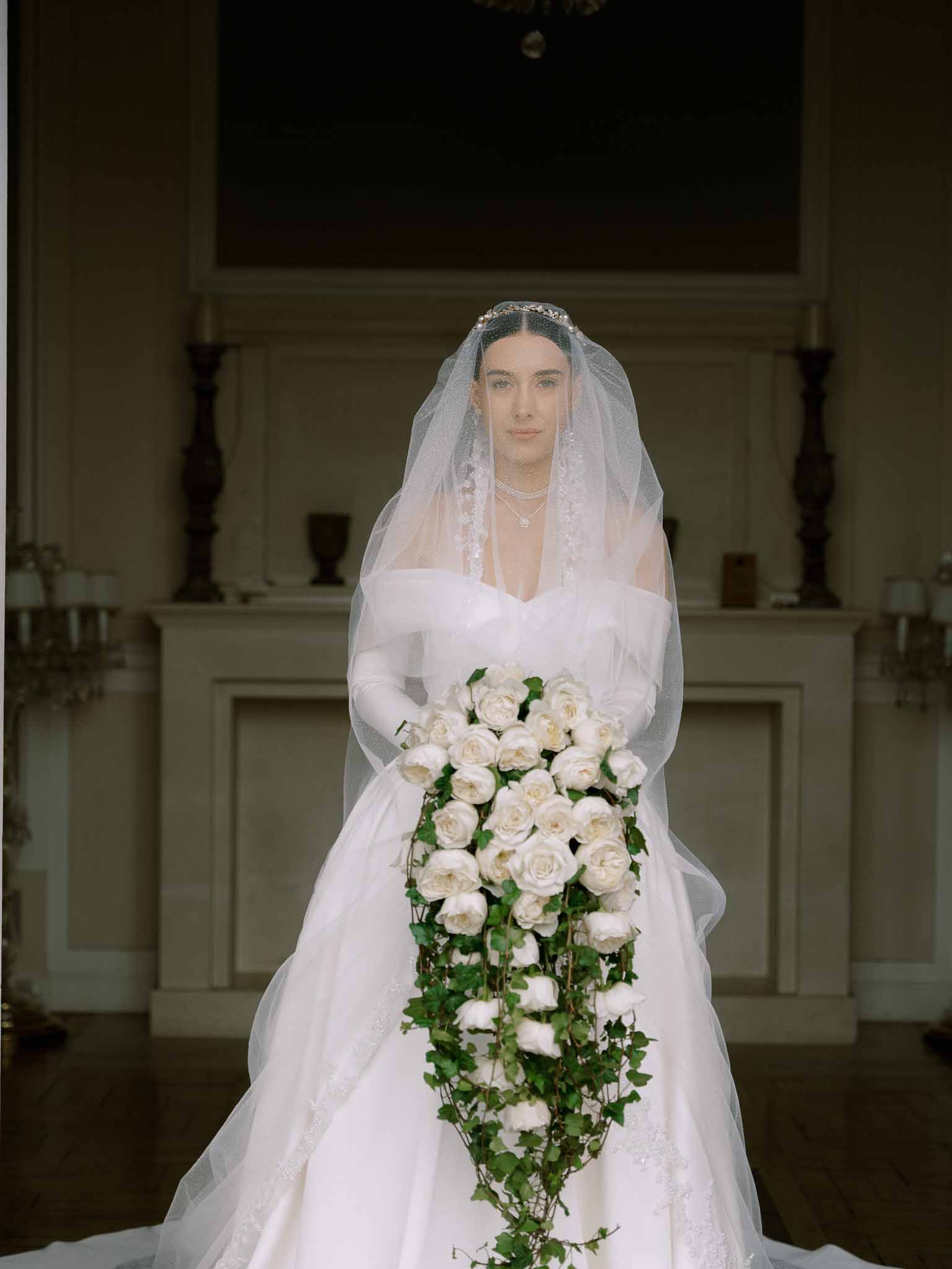 Bride in off-shoulder ivory ballgown with satin gloves holding cascading rose bouquet with ivy before white fireplace